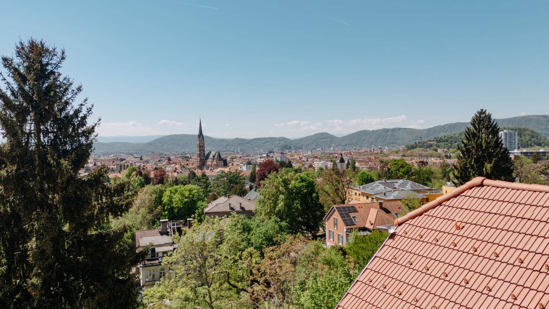 Panoramablick über die Stadt mit Kirchturm, grünen Hügeln und Bergen im Hintergrund.
