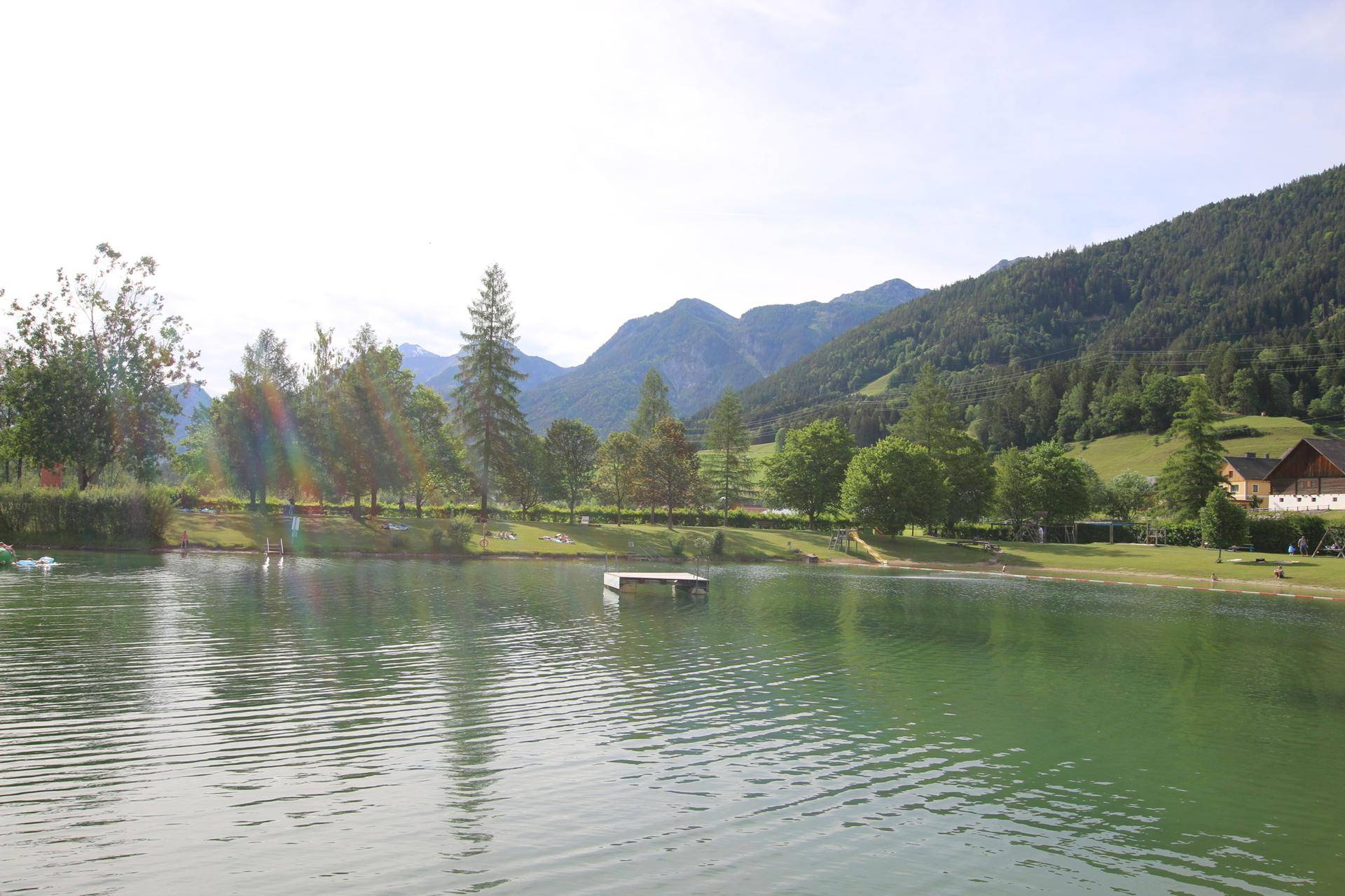 Ruhiger See mit einer Badeplattform, umgeben von Bäumen und einer malerischen Bergkulisse unter klarem Himmel.