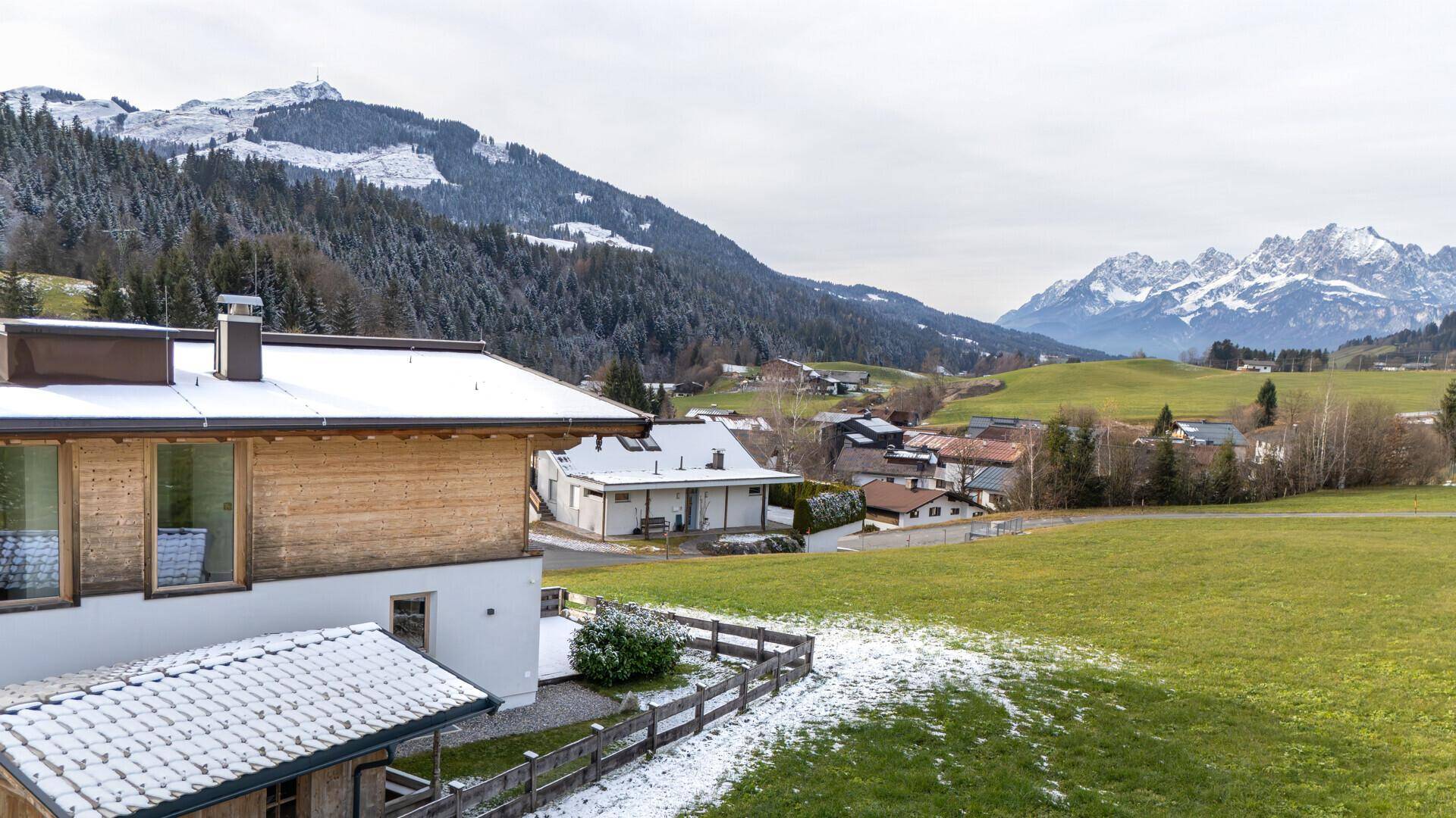 Weitläufige Landschaft mit schneebedeckten Bergen und grünen Hügeln, teilweise Blick auf ein Gebäude.