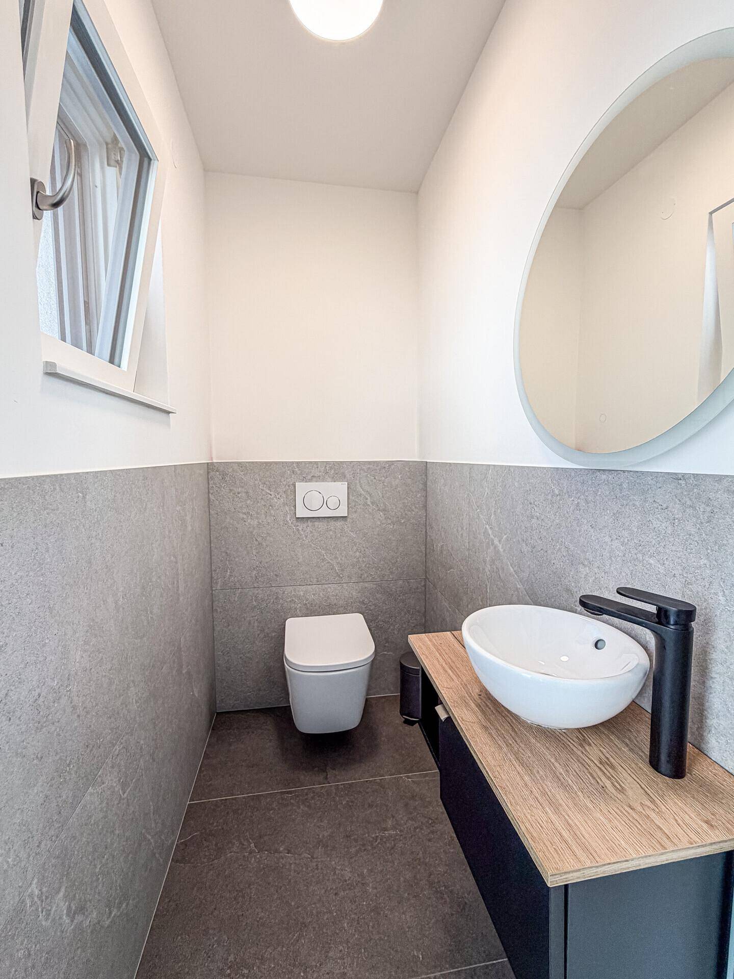 Modernes Gäste-WC mit grauen Fliesen, wandhängendem WC und Fenster.