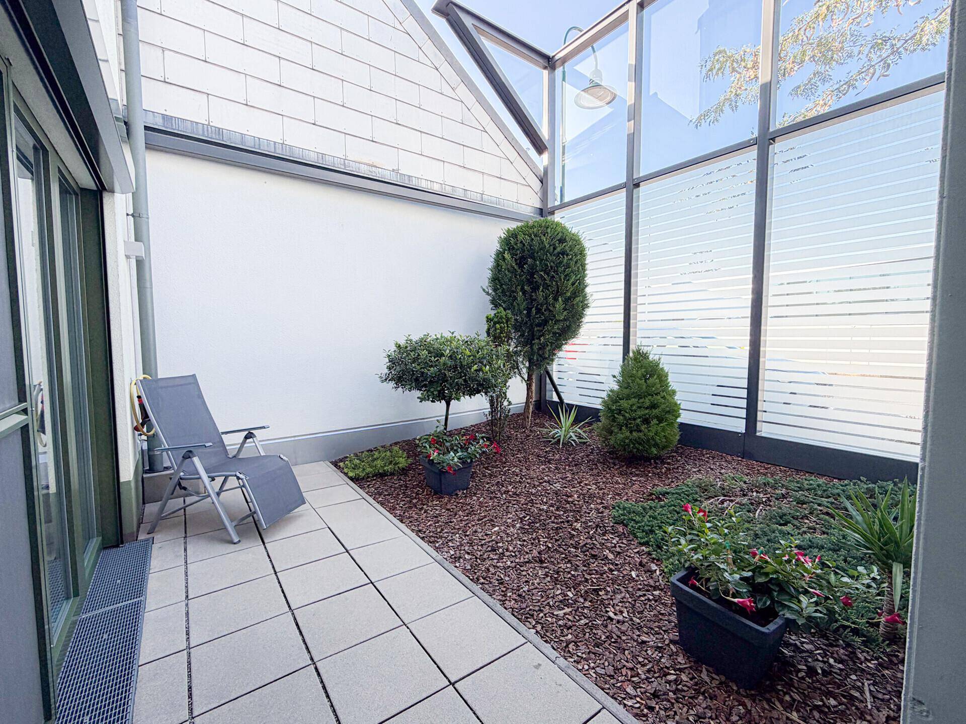Ruhige Terrasse mit Liegestuhl und bepflanztem Beet, umgeben von modernen Glaswänden.