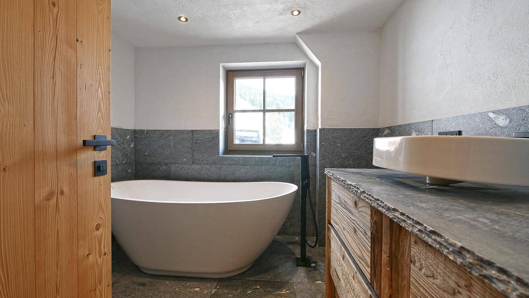 Modernes Badezimmer mit freistehender Badewanne, Natursteinfliesen und Holzwaschtisch unter einem Fenster.