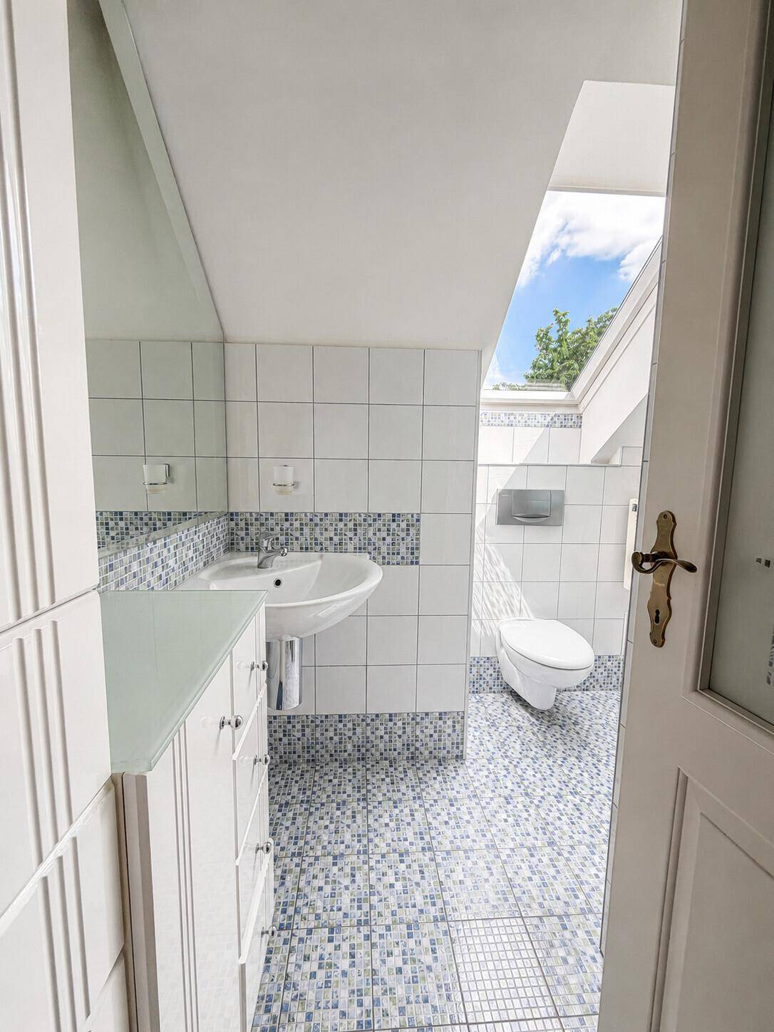 Helles Badezimmer im Dachgeschoss mit Dachfenster, weißen Fliesen und blauen Mosaikakzenten.