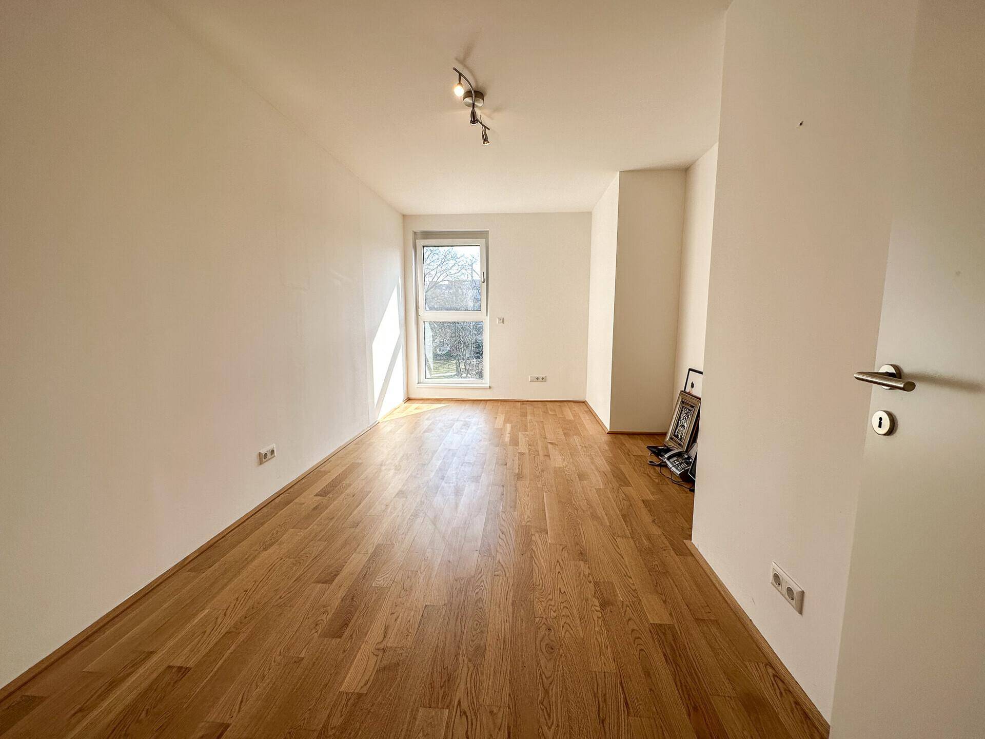 Helles Schlafzimmer mit Holzboden und großem Fenster, das viel Tageslicht hereinlässt.