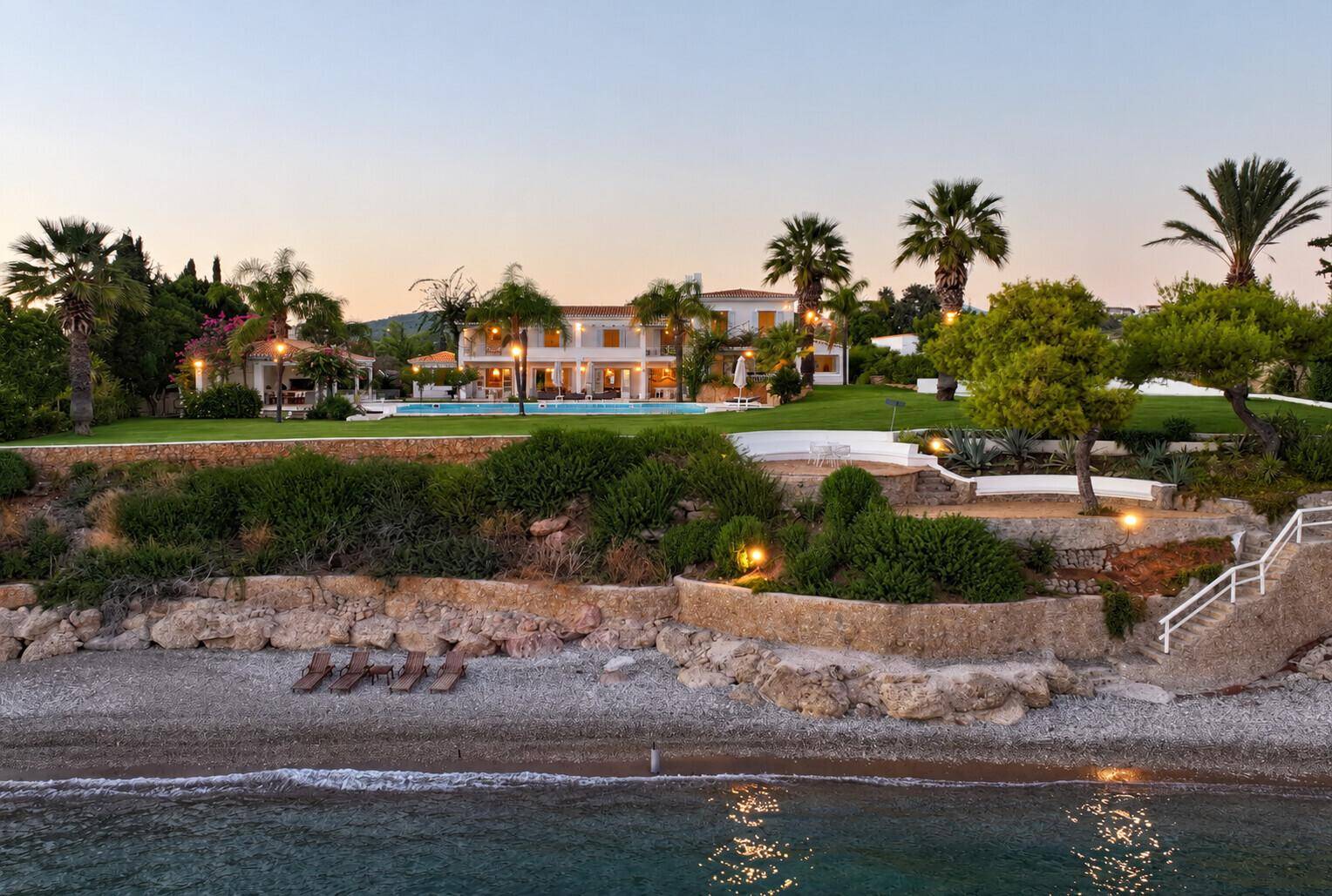 Luxuriöse Villa am Meer bei Sonnenuntergang, mit beleuchtetem Garten und direktem Strandzugang.