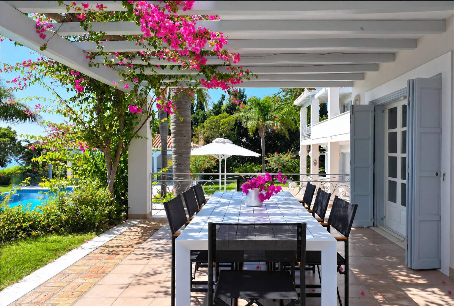 Überdachte Terrasse mit großem Esstisch, blühender Bougainvillea und Blick auf den Garten und Pool.