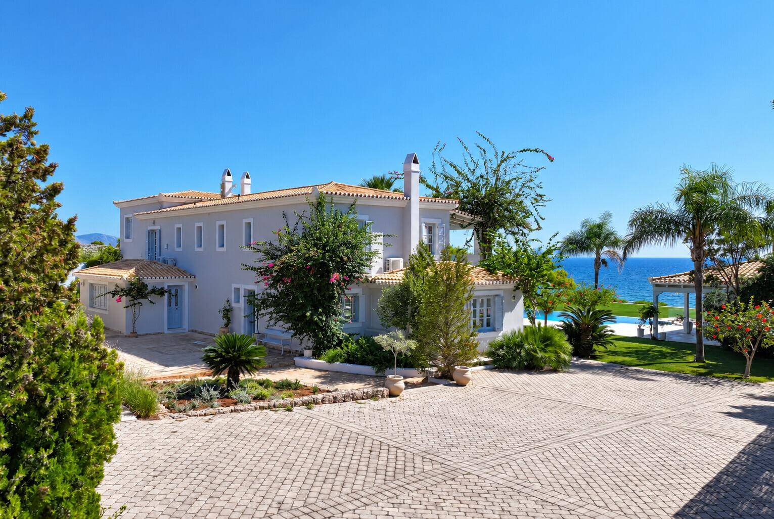 Elegante Villa mit großem gepflastertem Vorplatz, umgeben von mediterraner Vegetation und Meerblick.
