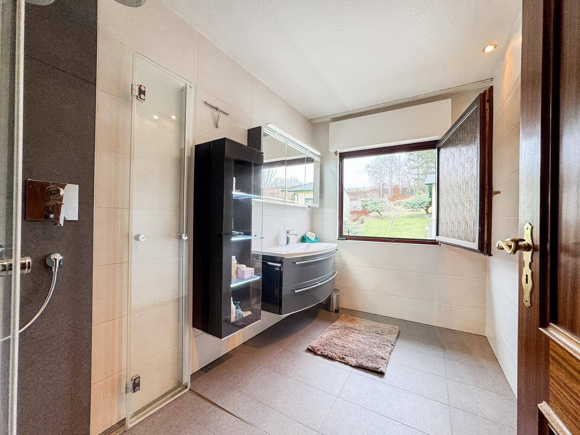 Modernes Badezimmer mit ebenerdiger Dusche, Hängeschrank und Fenster mit Blick ins Grüne.