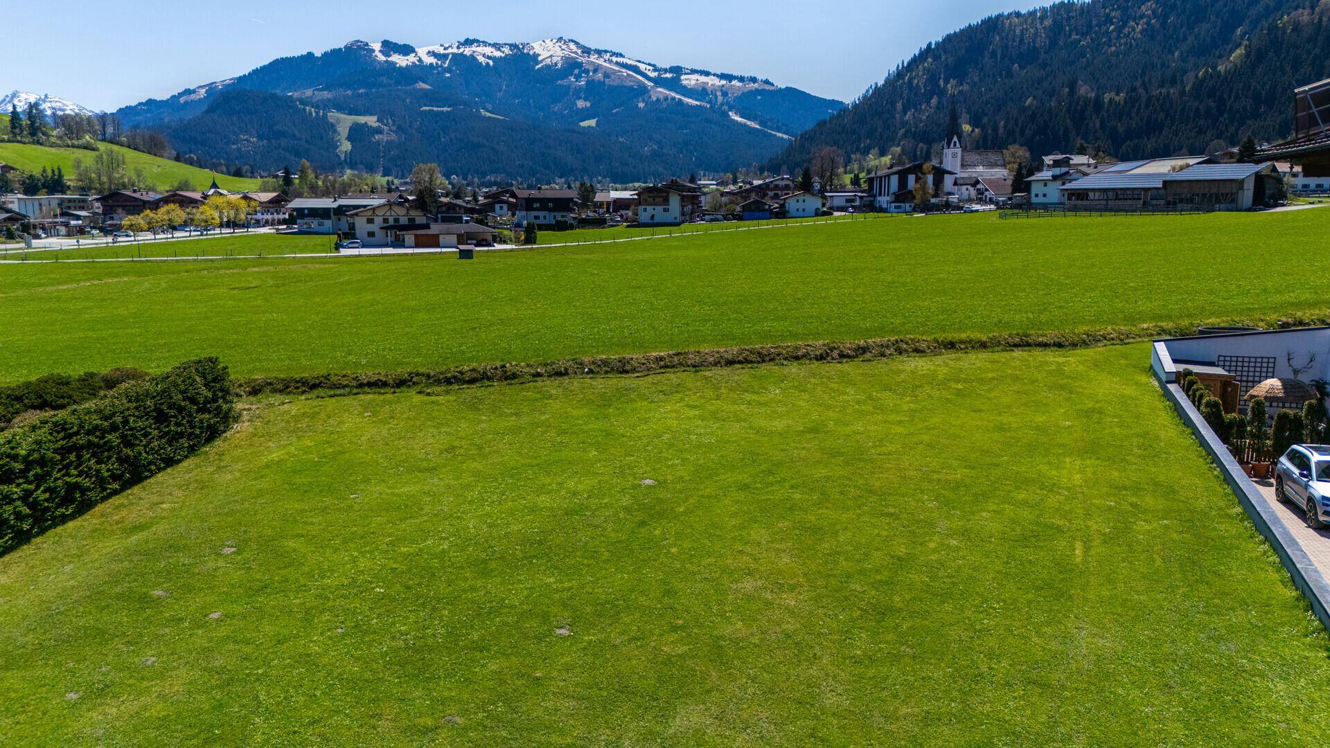 Weitläufige grüne Wiesenfläche mit einem Dorf und schneebedeckten Bergen in der Ferne unter blauem Himmel.