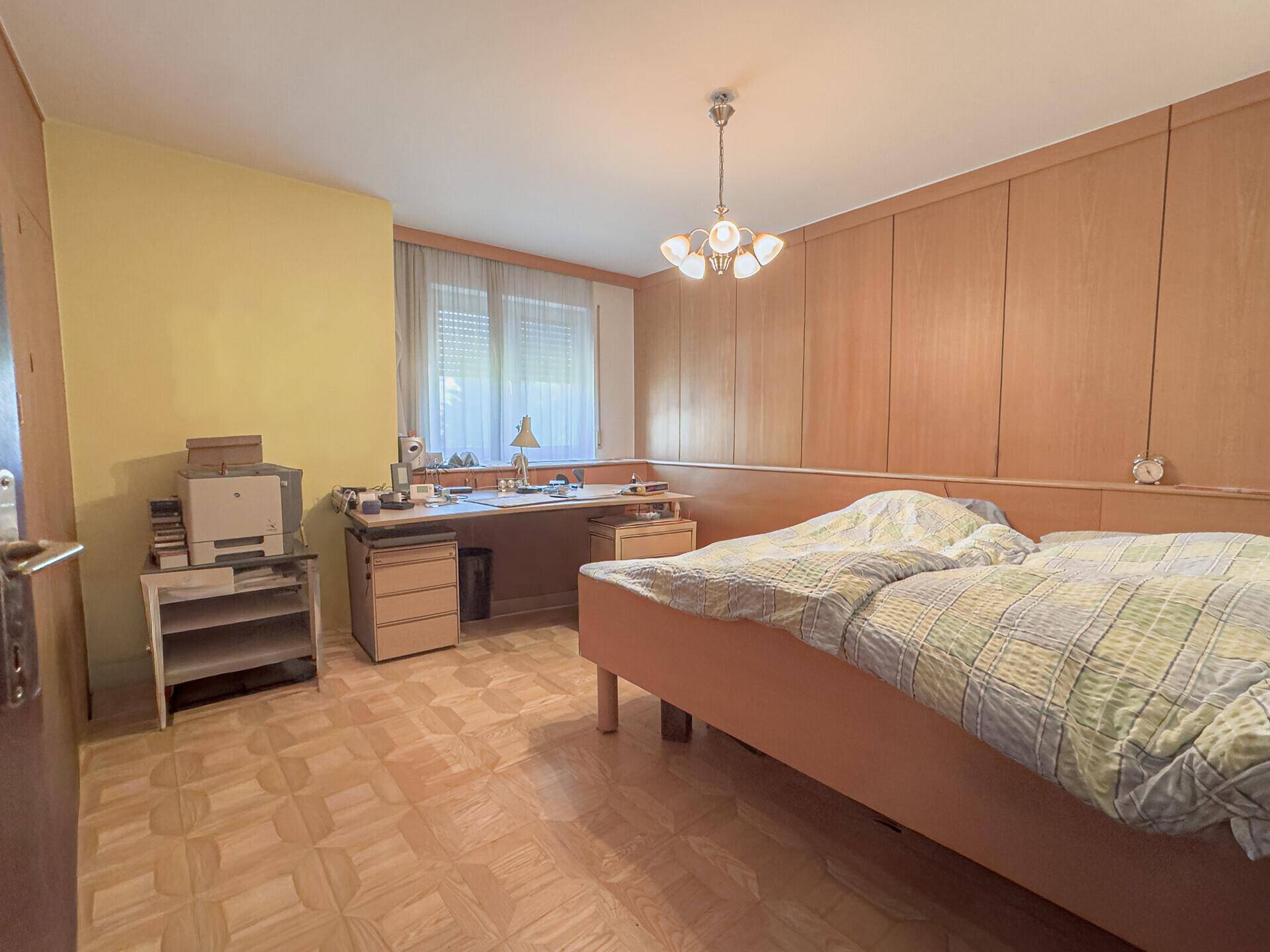 Schlafzimmer mit Holzvertäfelung, Parkettboden, Bett und einem Arbeitsbereich am Fenster.