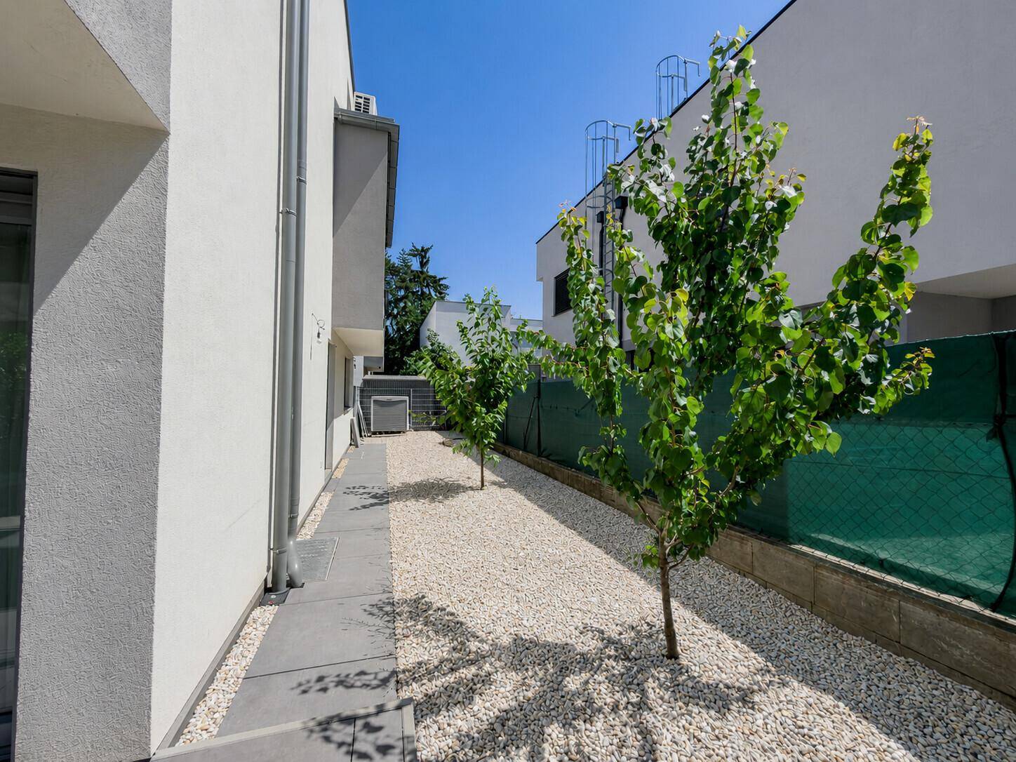 Seitenansicht des modernen Hauses mit Kiesweg und jungen Bäumen unter blauem Himmel.