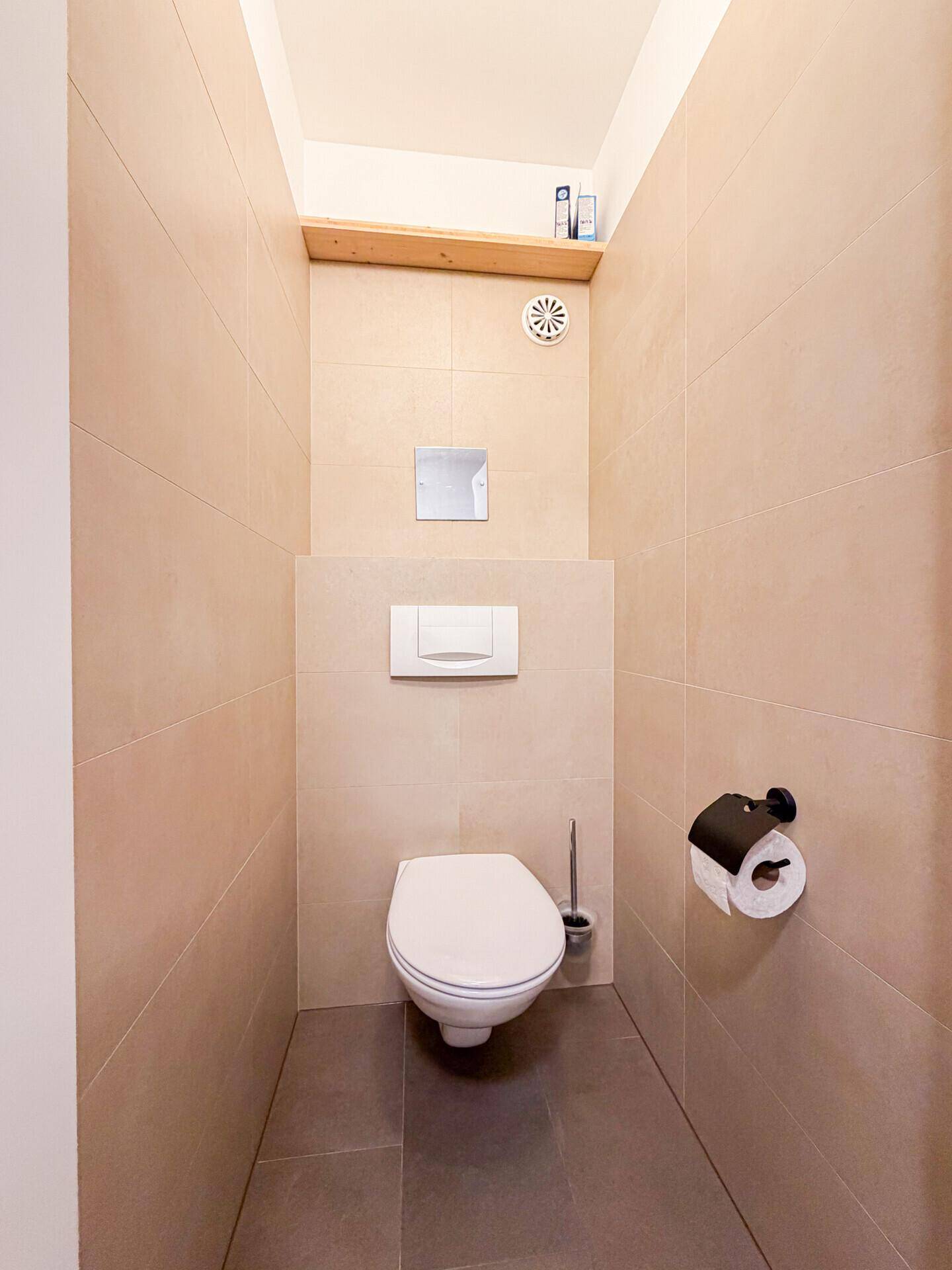 Separates WC mit wandhängender Toilette und beigefarbenen Wandfliesen.