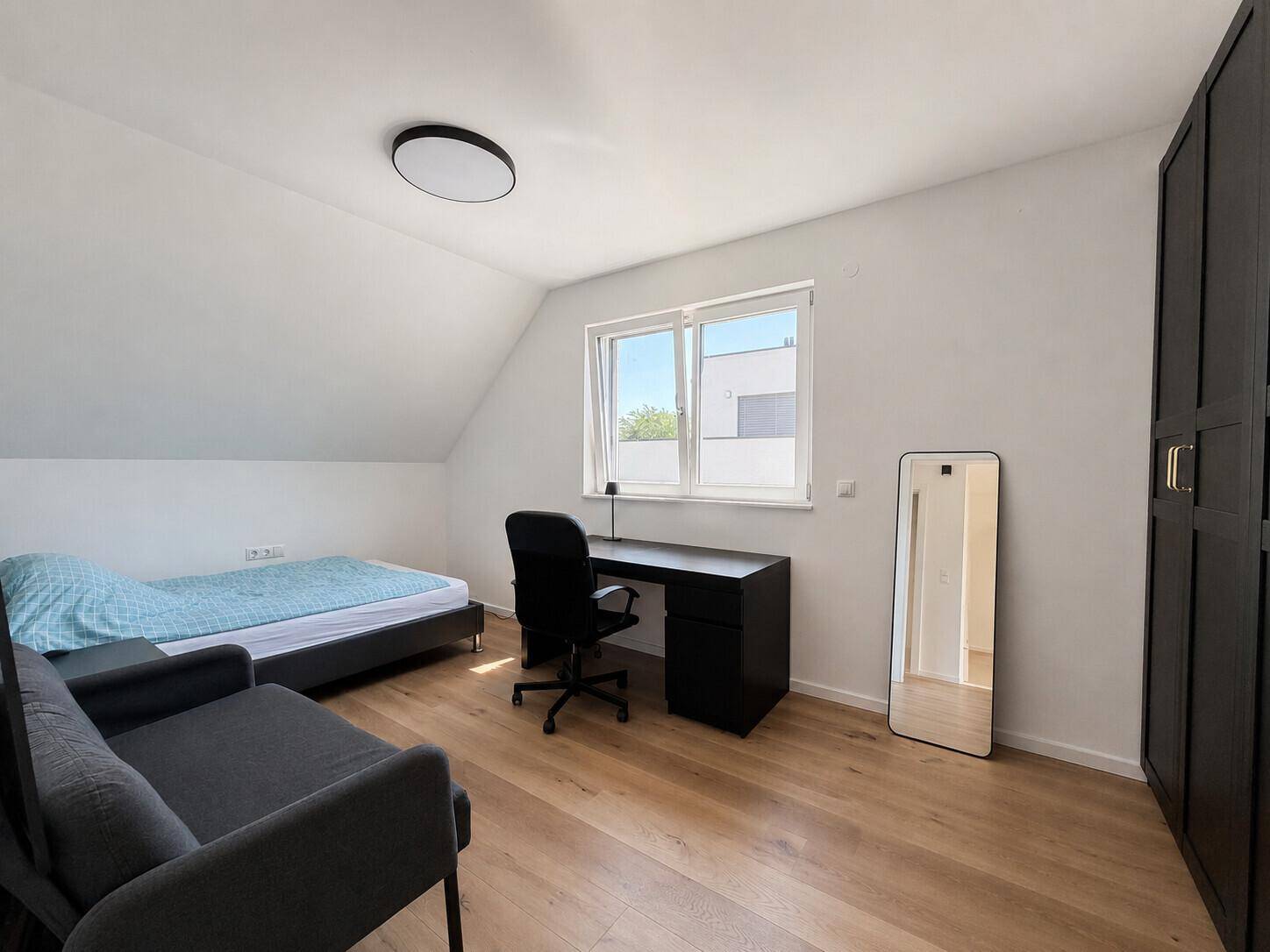 Helles Zimmer im Dachgeschoss mit Holzboden, Schreibtisch und Fenster.