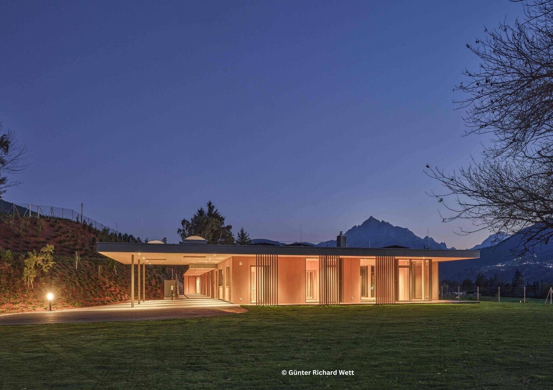 Moderne Villa bei Nacht mit beleuchtetem Carport und beeindruckender Bergkulisse im Hintergrund.