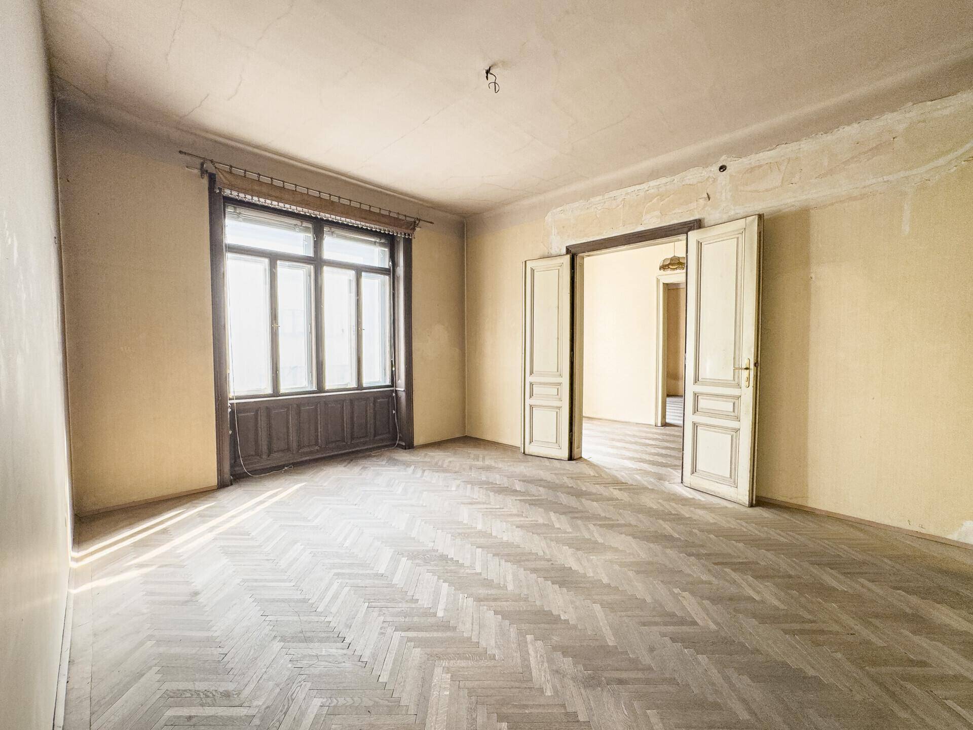 Leerer Wohnraum mit Fischgrätparkett, hohen Decken und einem großen Fenster, das viel Licht hereinlässt.