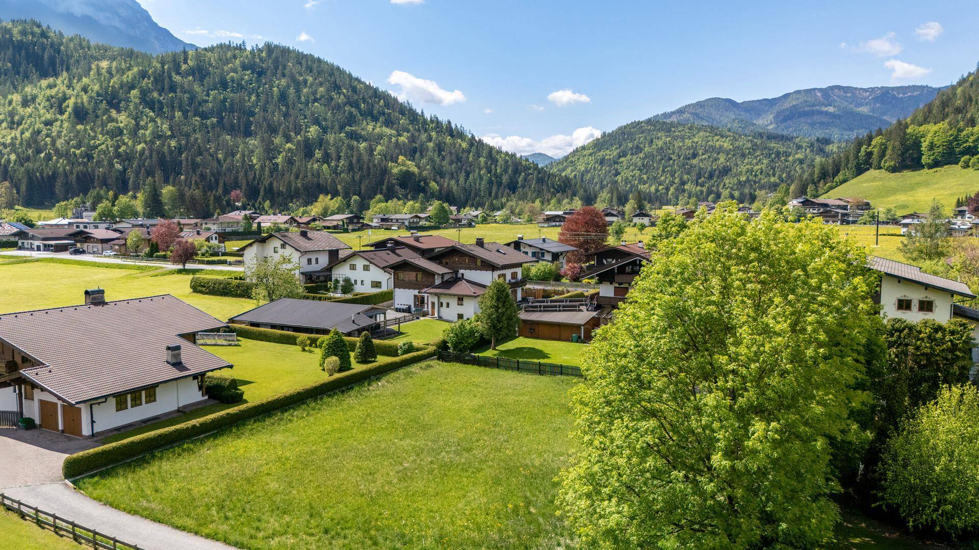 Wohngebiet in einem grünen Tal, umgeben von bewaldeten Bergen unter strahlend blauem Himmel.