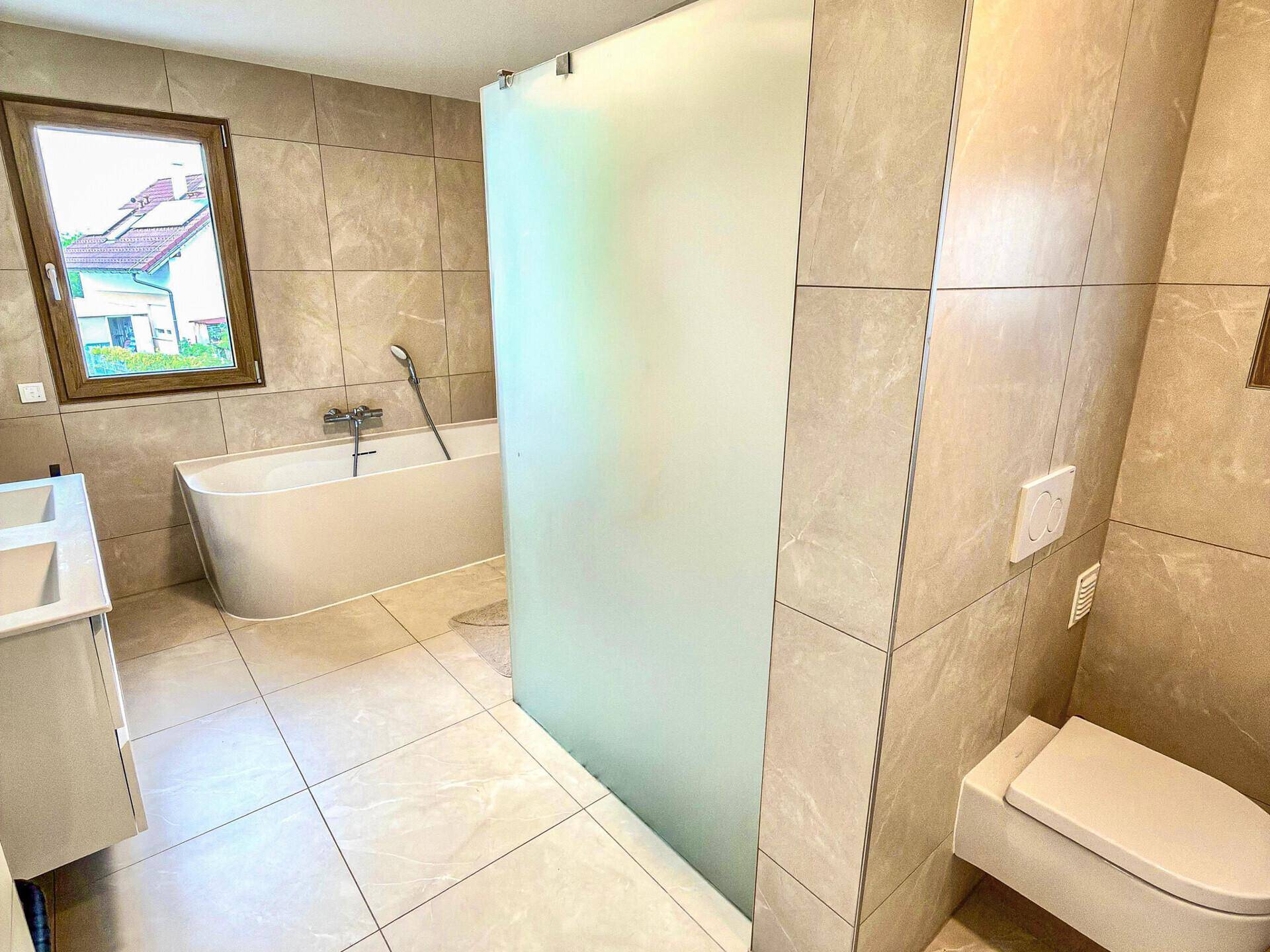 Modernes Badezimmer mit Badewanne, Dusche und Wand-WC, hell gefliest und mit Fenster für Tageslicht.