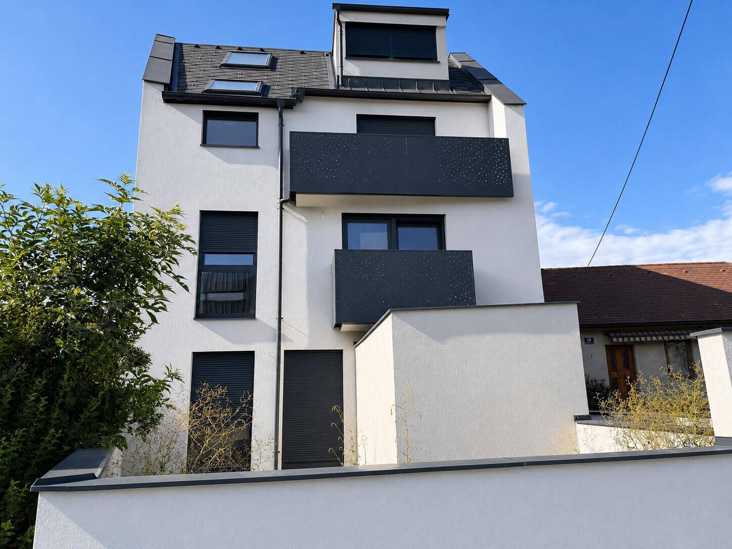 Moderne weiße Fassade eines Neubaus mit dunklen Balkonen und Dachfenstern unter blauem Himmel.