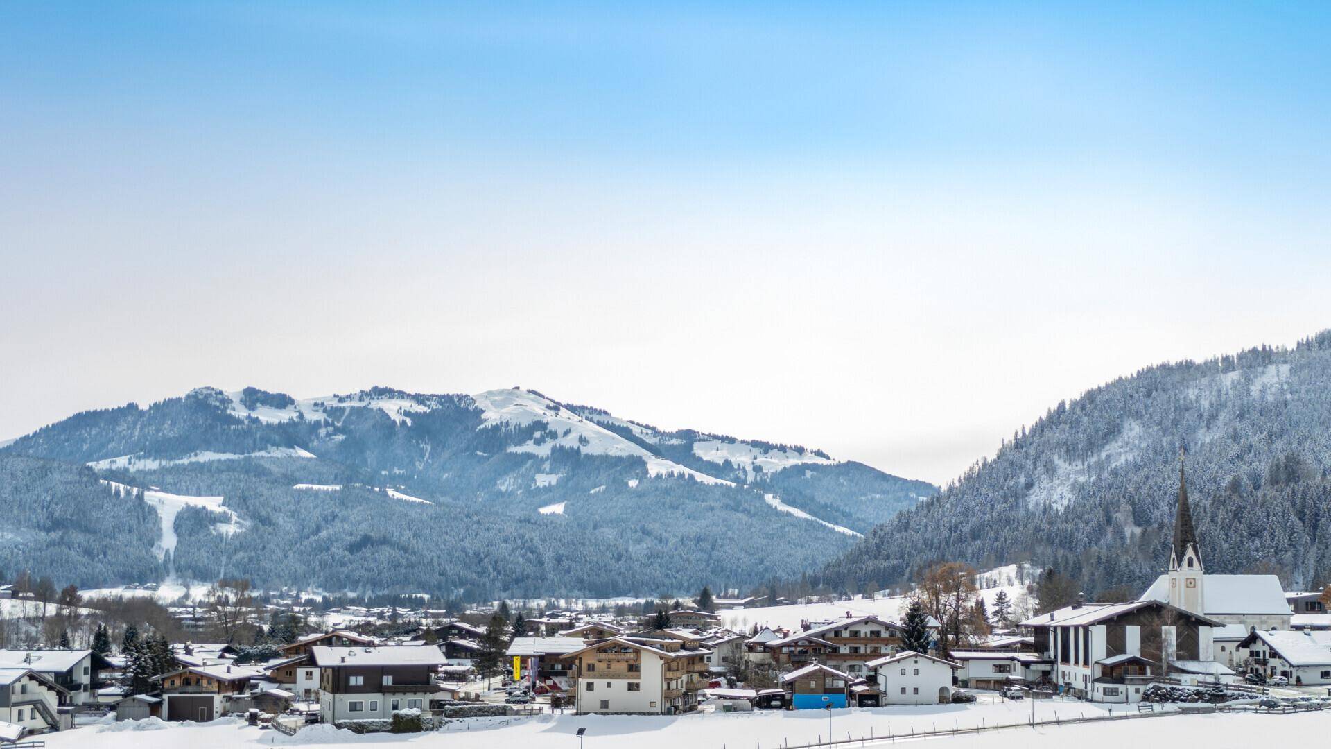 Panoramablick auf ein idyllisches, verschneites Dorf mit markanter Kirche und umliegenden Bergen.