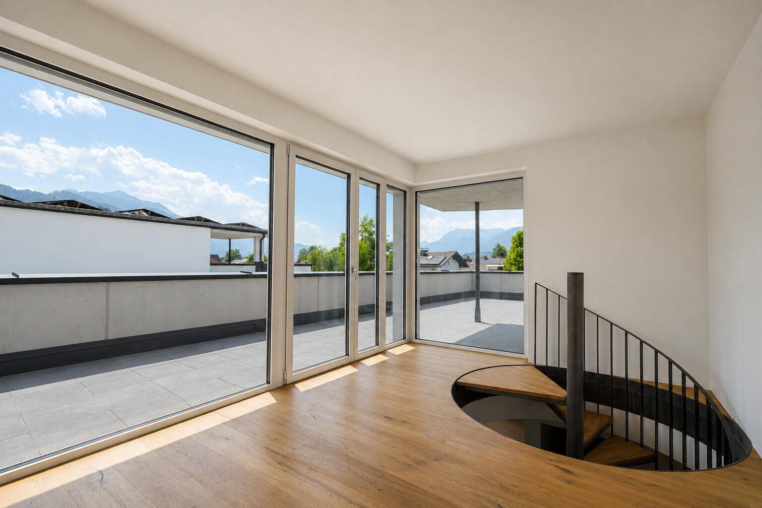 Heller Raum mit Holzboden, Wendeltreppe und großen Fensterfronten mit Bergblick.
