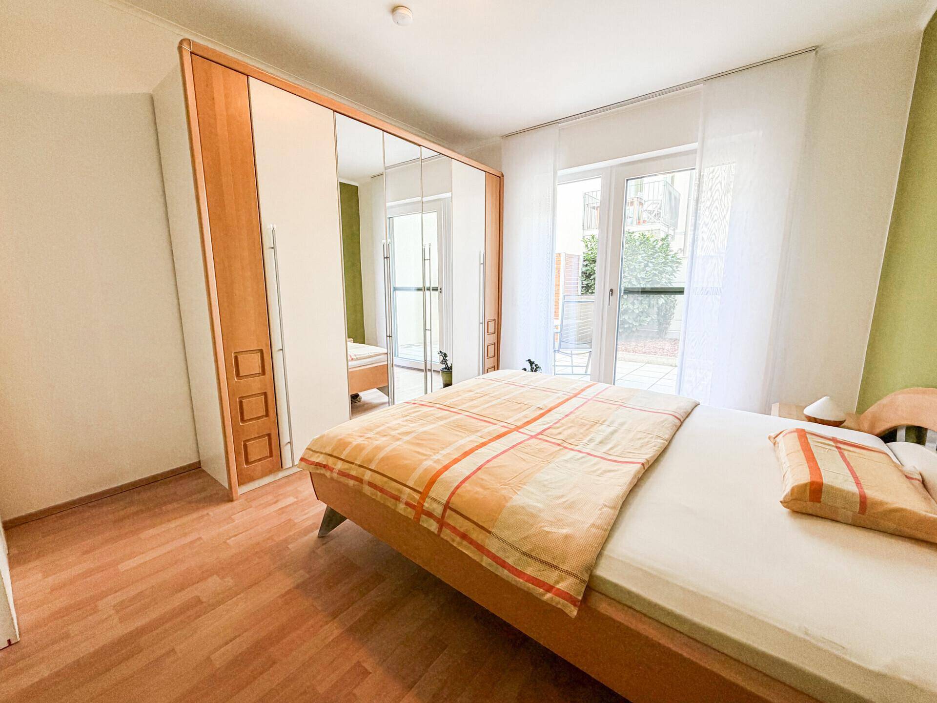 Schlafzimmer mit großem Spiegelschrank, Holzbett und hellem Zugang zum Außenbereich.