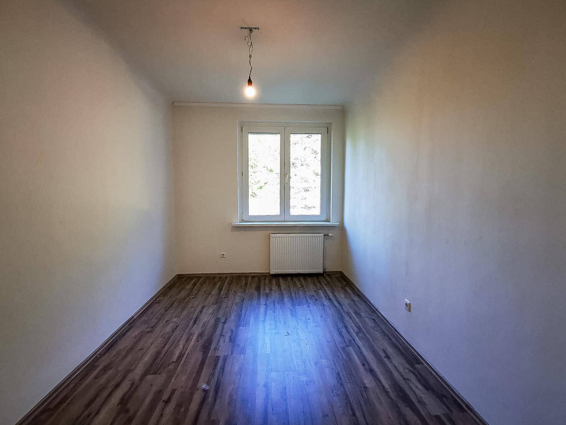 Schmales, helles Zimmer mit Laminatboden und einem Fenster mit Blick ins Grüne.