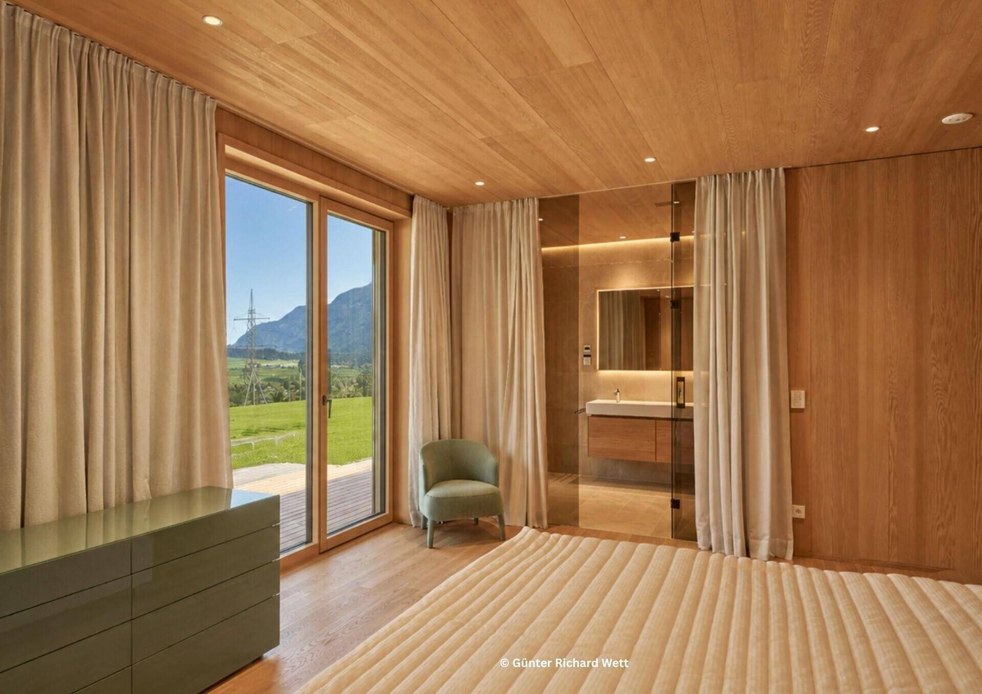 Helles Schlafzimmer mit Holzboden, großen Fenstern und integriertem Bad mit Bergblick.