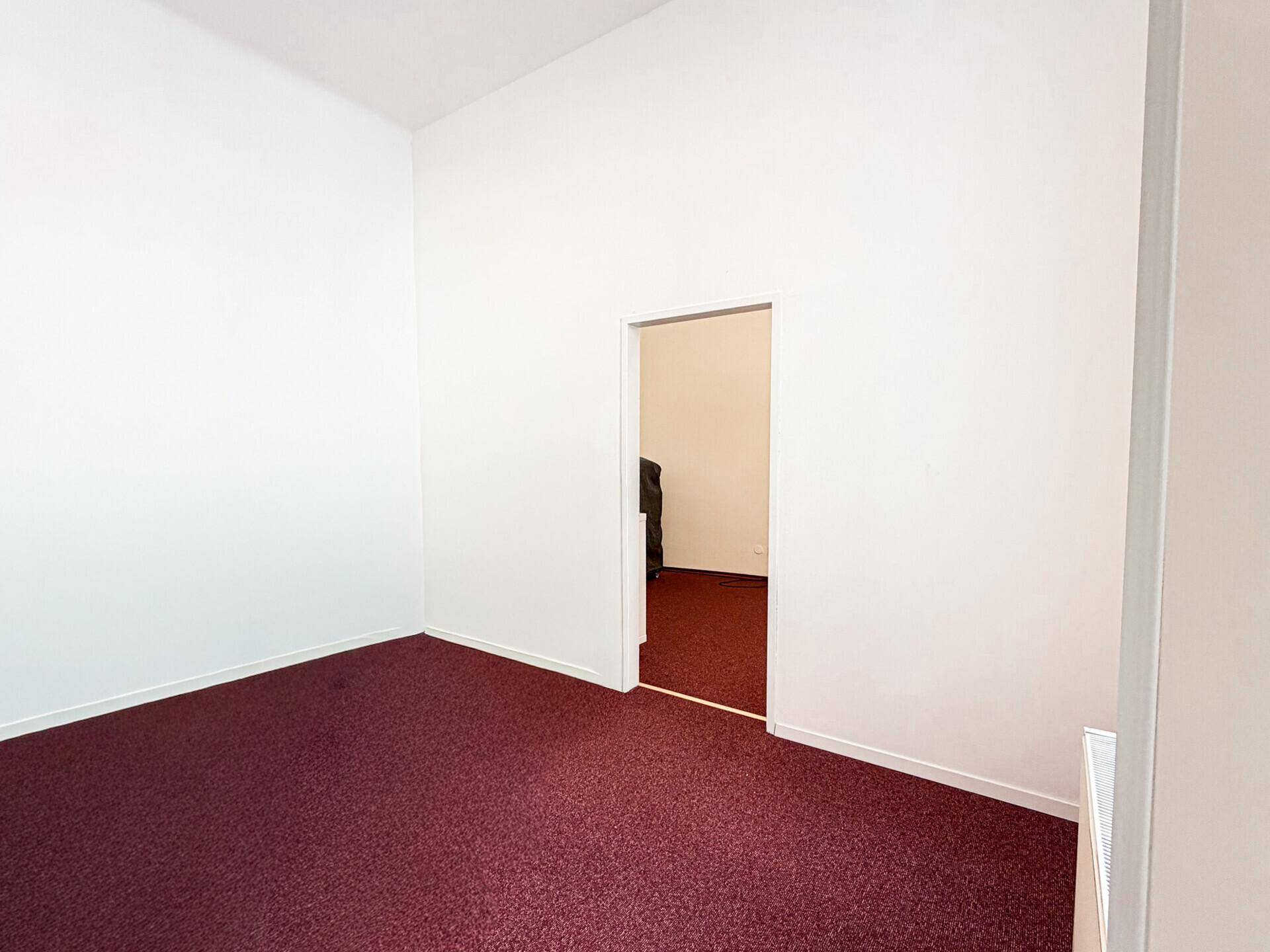 Einfaches Zimmer mit rotem Teppichboden und einer Tür, die zu einem weiteren Raum führt.