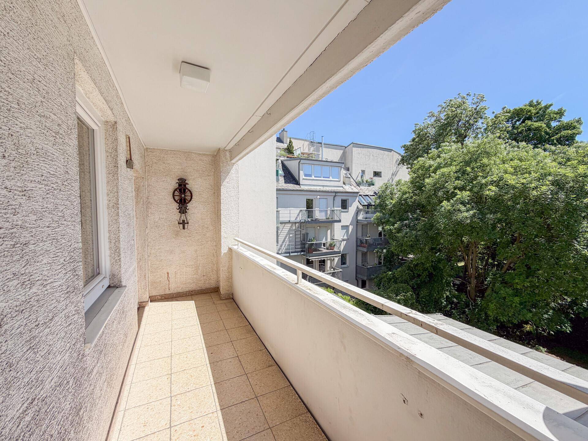 Offener Balkon mit Fliesenboden und Blick auf die umliegenden Gebäude und Bäume.