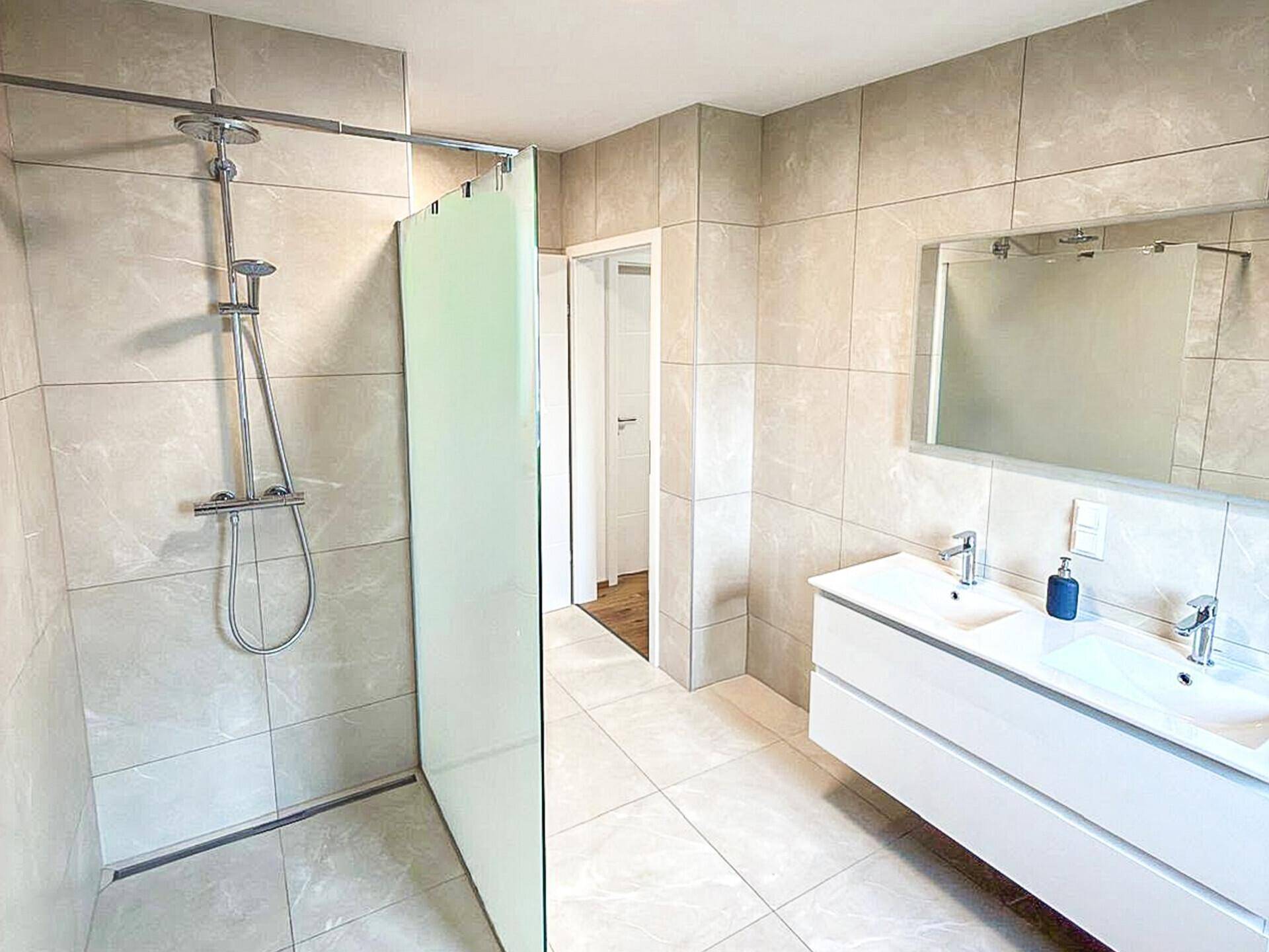 Modernes Badezimmer mit Doppelwaschbecken, begehbarer Dusche und großen Fliesen, hell und geräumig.