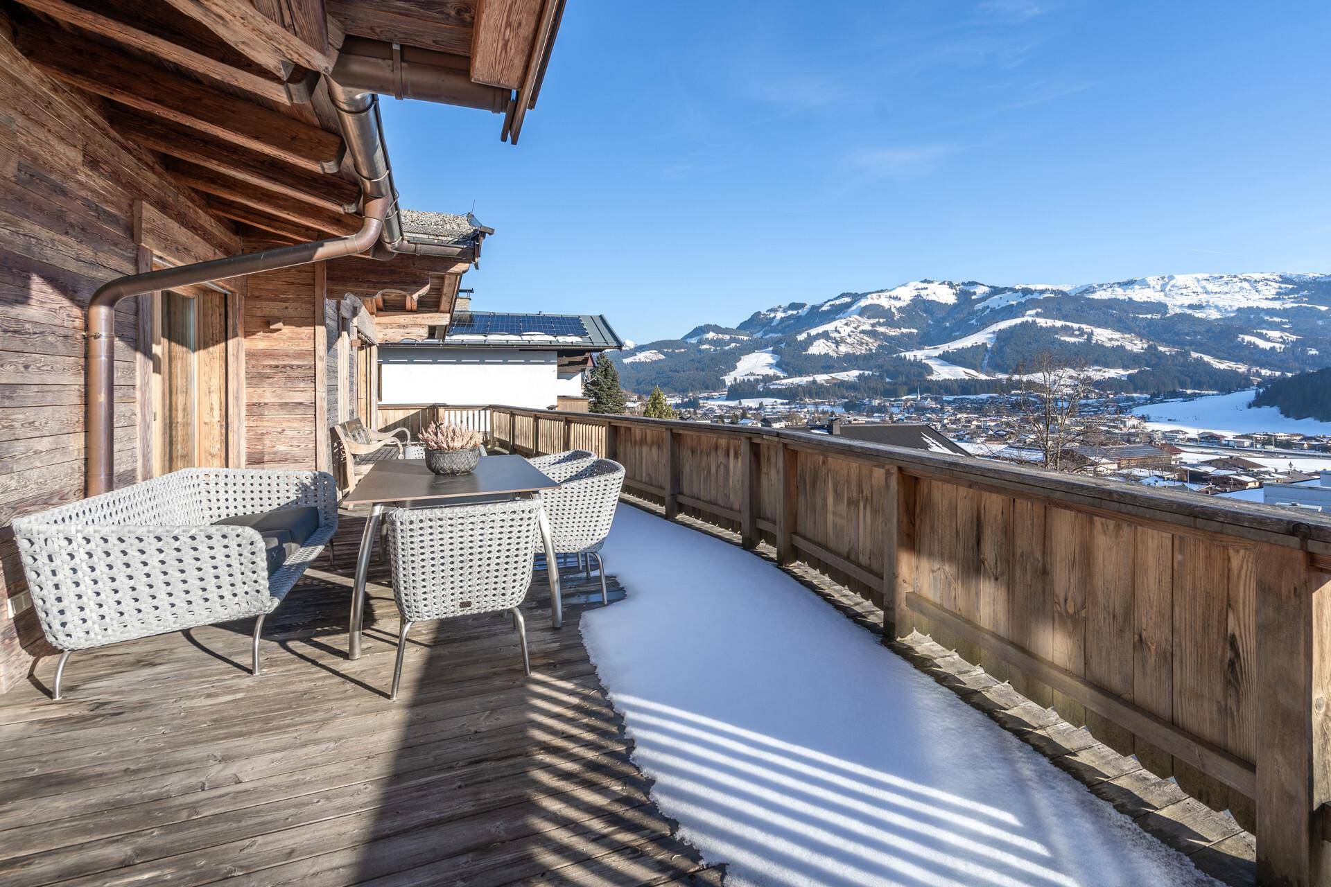 Sonniger Holzbalkon mit modernen Outdoor-Möbeln und weitem Blick über das verschneite Tal und die Berge.