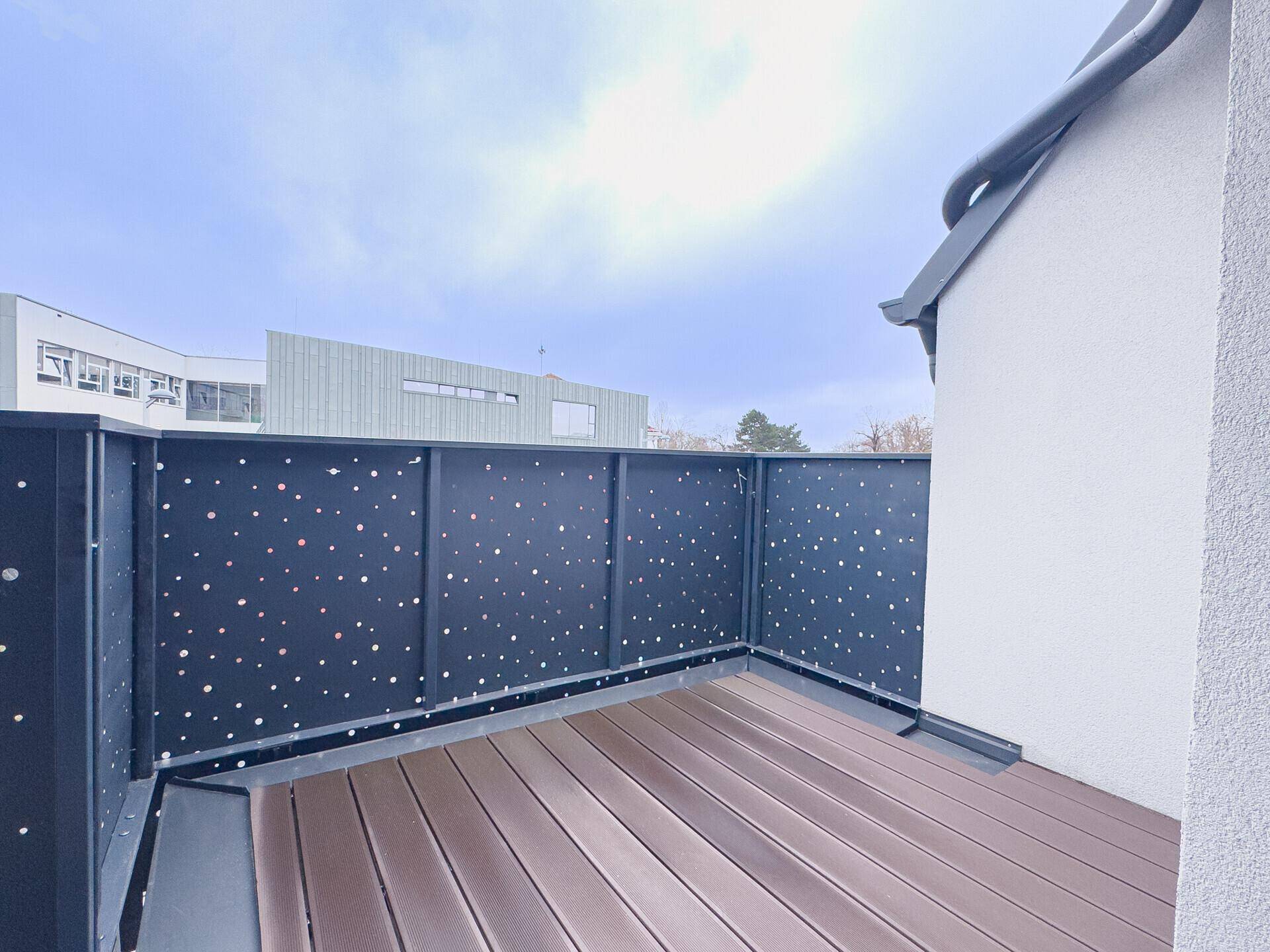 Moderner Balkon mit dunklem, gemustertem Geländer und Holzdielenboden unter offenem Himmel.