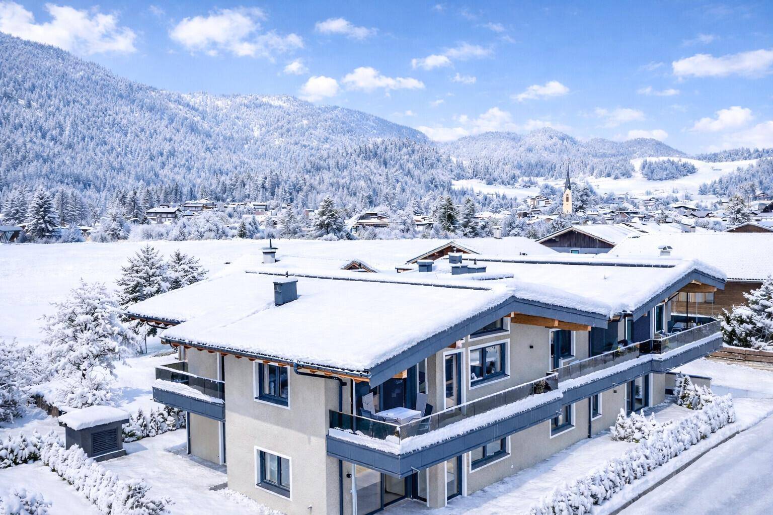 Winterliche Luftaufnahme eines modernen Mehrfamilienhauses mit schneebedeckten Dächern und Bergblick.