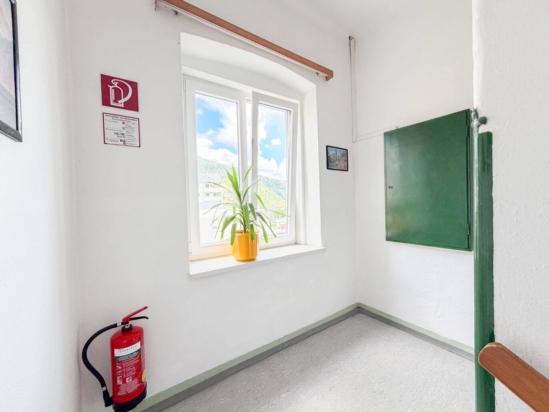 Heller Treppenabsatz mit Fenster, Blick auf die Berge und einem Feuerlöscher.