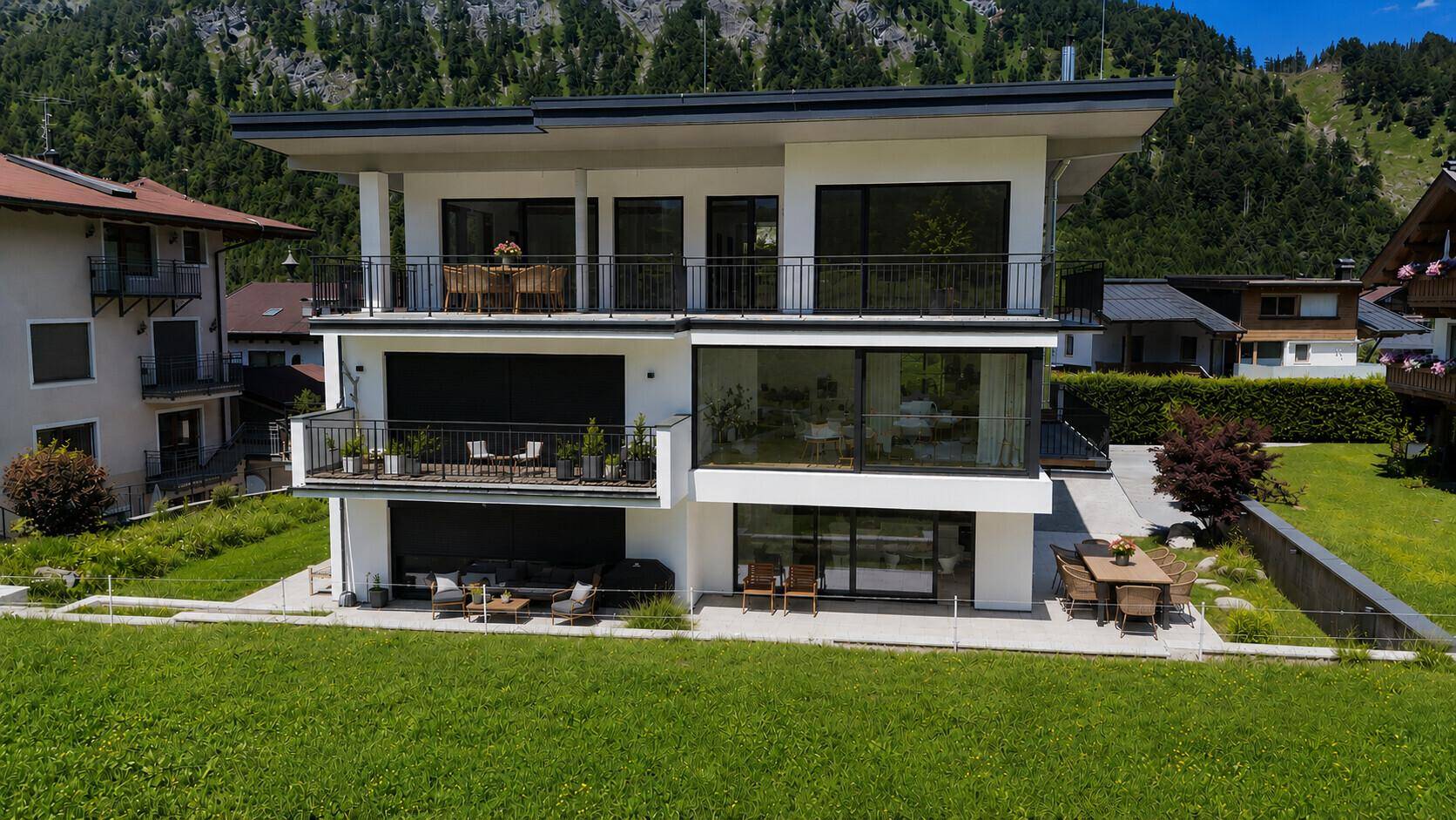 Moderne Villa mit mehreren Balkonen und Terrassen, umgeben von grüner Landschaft und Bergen.