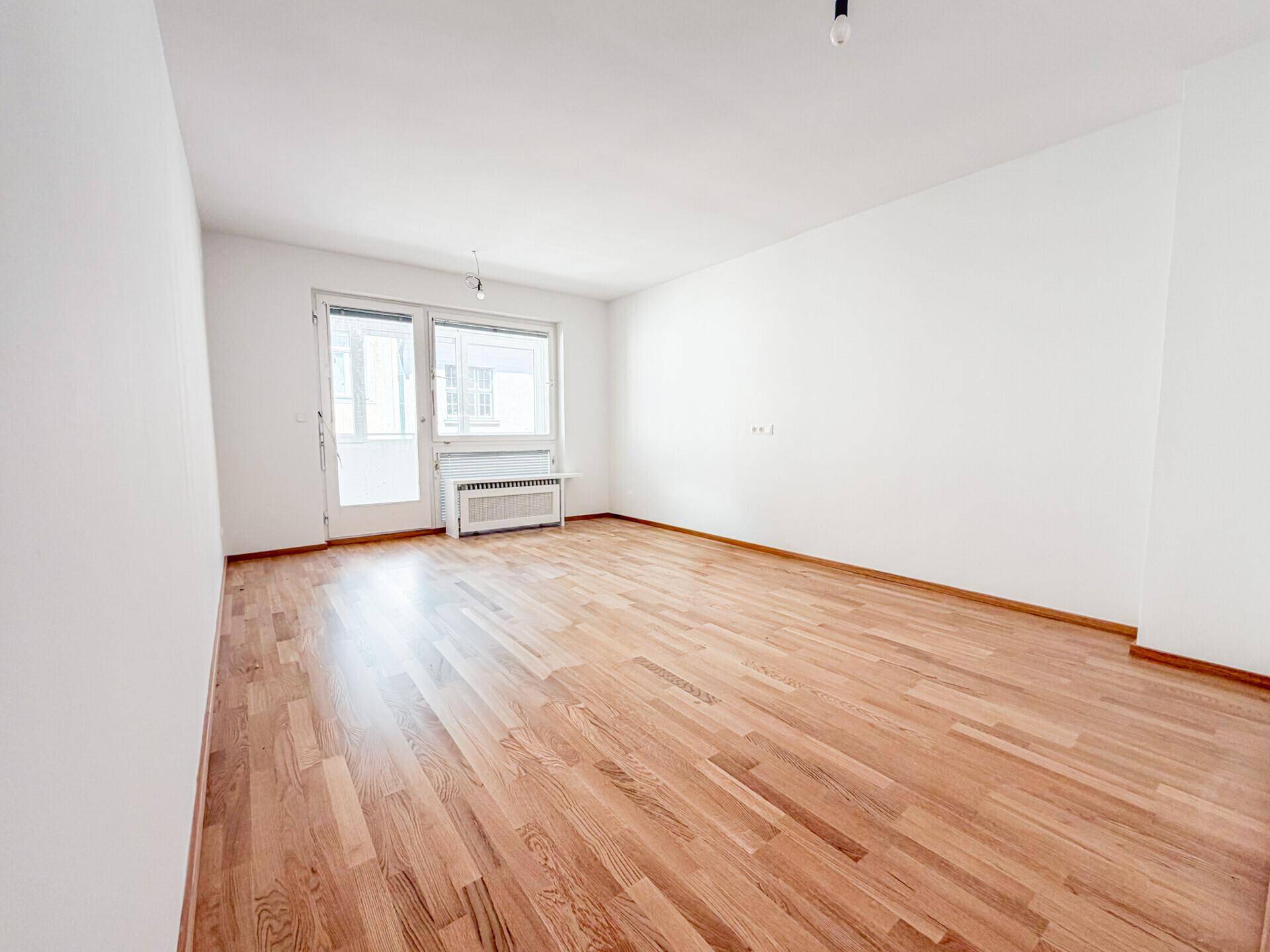 Geräumiges Wohnzimmer mit hellem Parkettboden, weißen Wänden und Zugang zu einem Balkon oder einer Loggia.