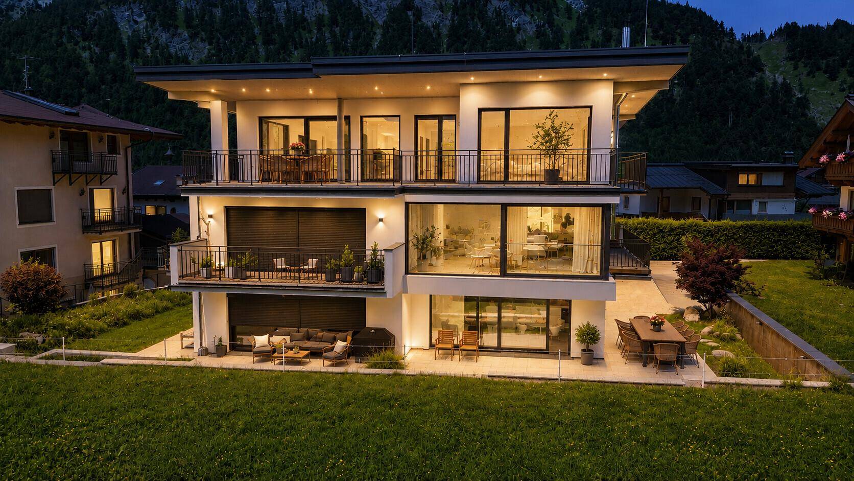 Moderne Villa bei Dämmerung, beleuchtet mit warmem Licht und Blick auf die umliegenden Berge.
