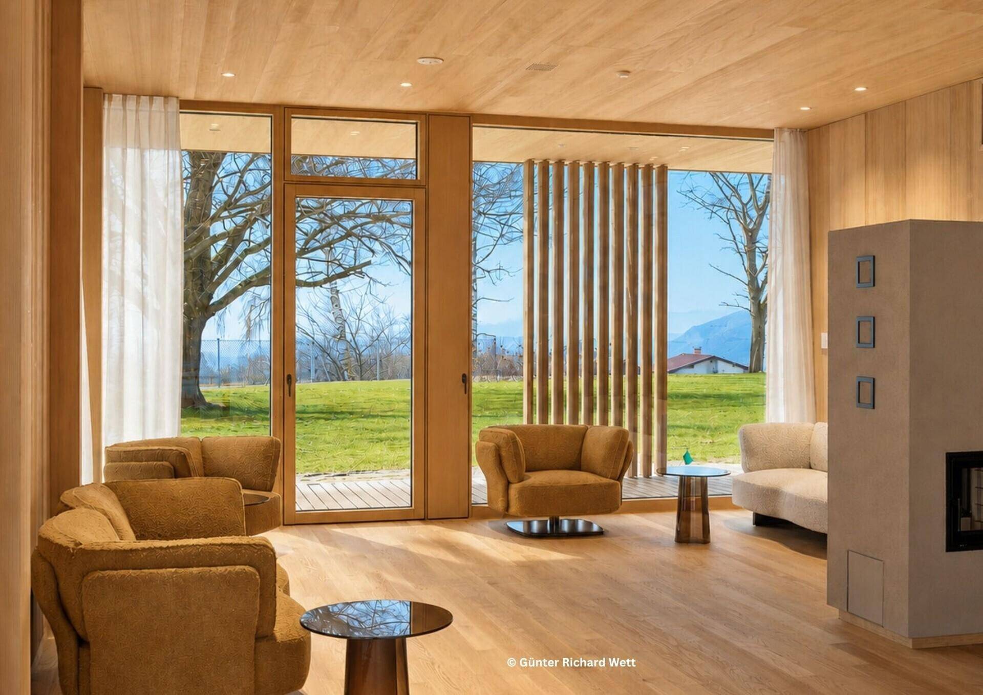 Lichtdurchflutetes Wohnzimmer mit Holzboden, modernen Sesseln und Kaminofen mit Blick ins Grüne.
