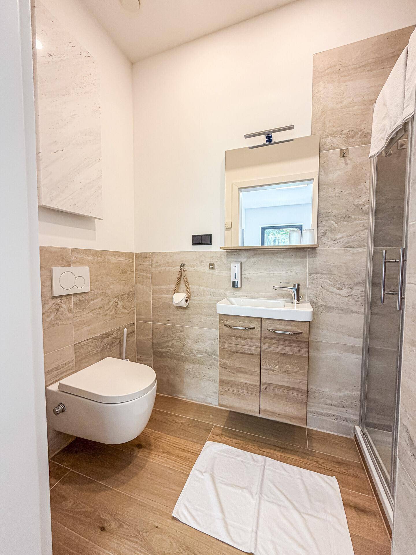 Modernes Gäste-WC mit Wand-WC, kleinem Waschbecken und Duschkabine in hellen Farbtönen.