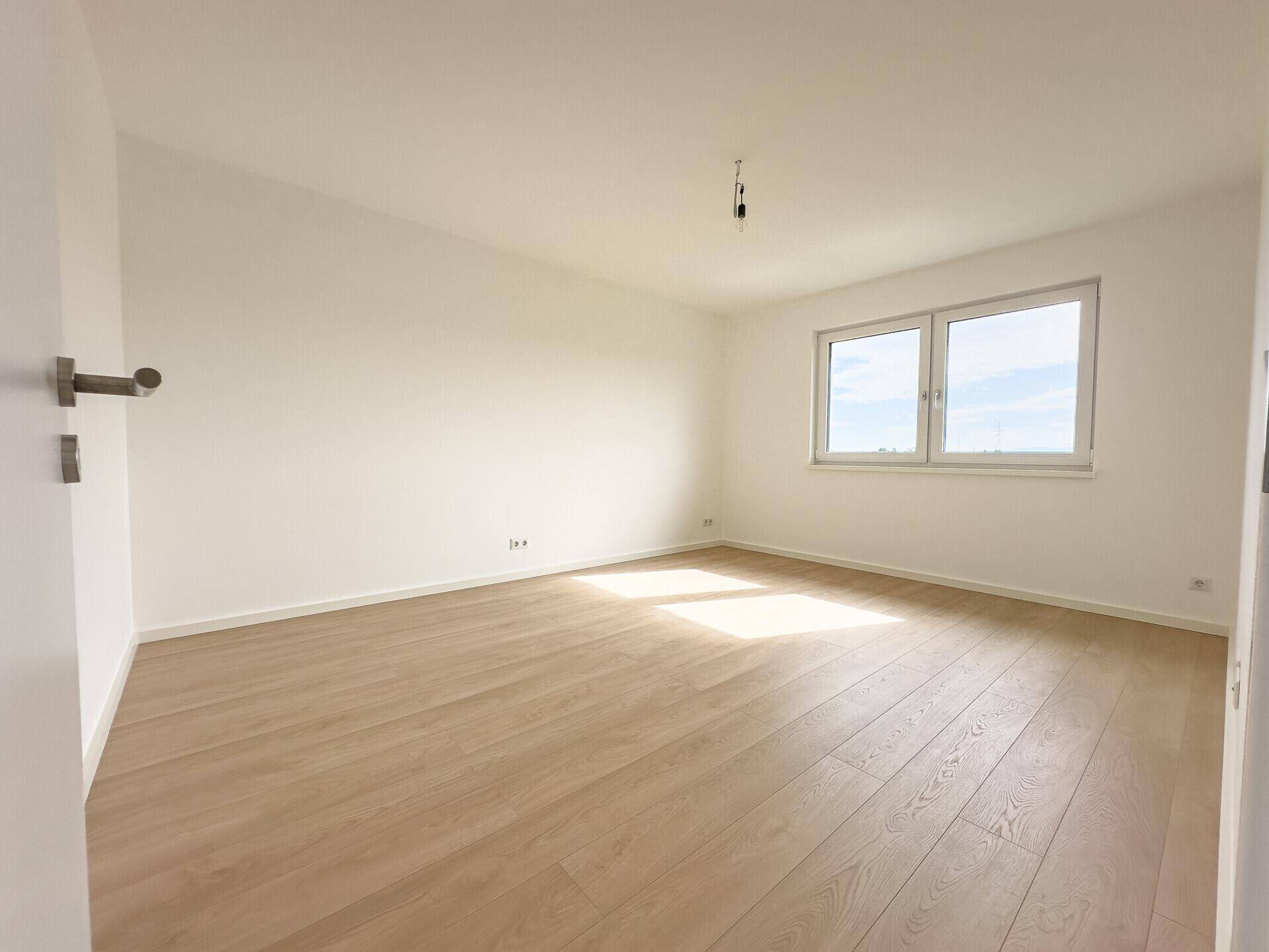 Geräumiges, helles Zimmer mit Holzboden und großen Fenstern, bietet viel Tageslicht.