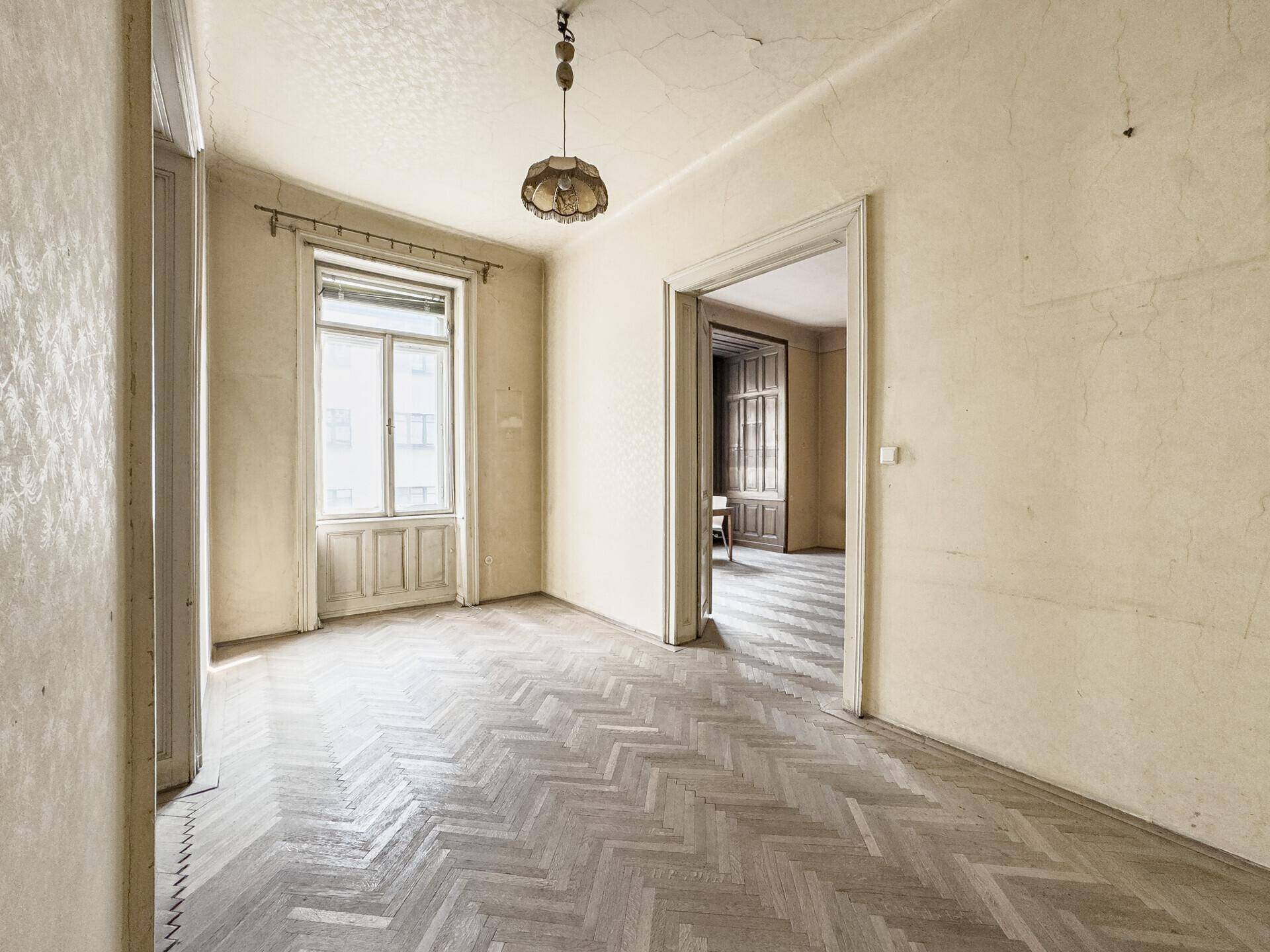 Leeres Zimmer mit Fischgrätparkett, hohen Decken und einem Fenster, das Tageslicht spendet.