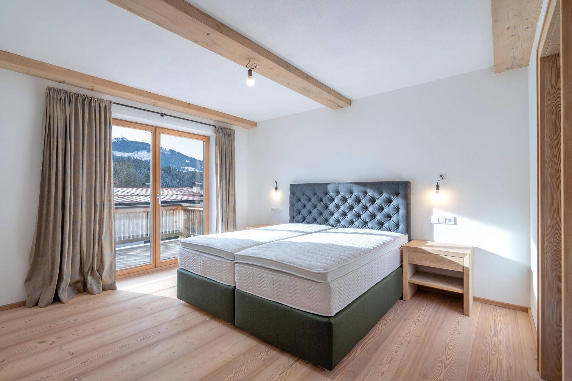 Helles Schlafzimmer mit Doppelbett, Holzbalken und Balkonzugang mit Blick auf die schneebedeckten Berge.