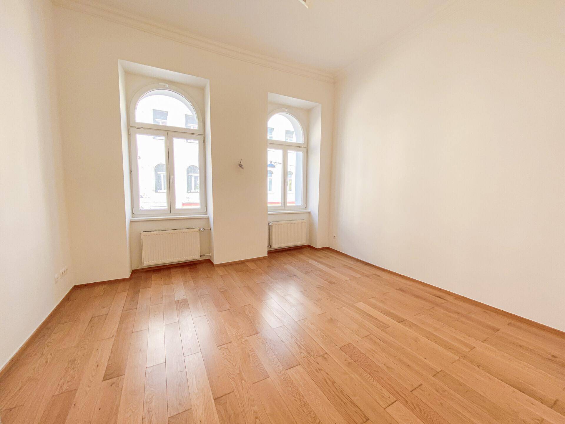 Geräumiges, helles Zimmer mit Holzboden, hohen Decken und zwei großen Fenstern.