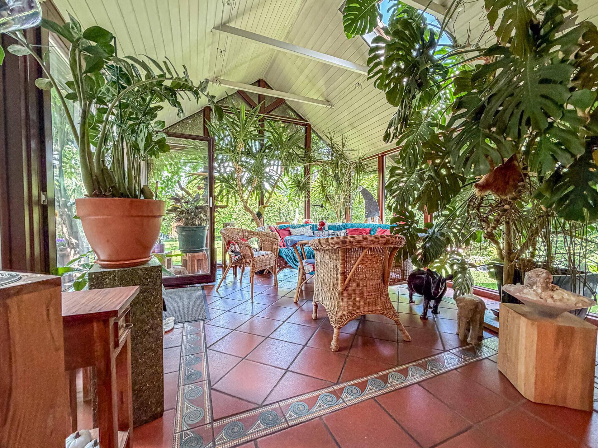 Heller Wintergarten mit vielen Pflanzen, Korbmöbeln und Terrakotta-Fliesenboden.