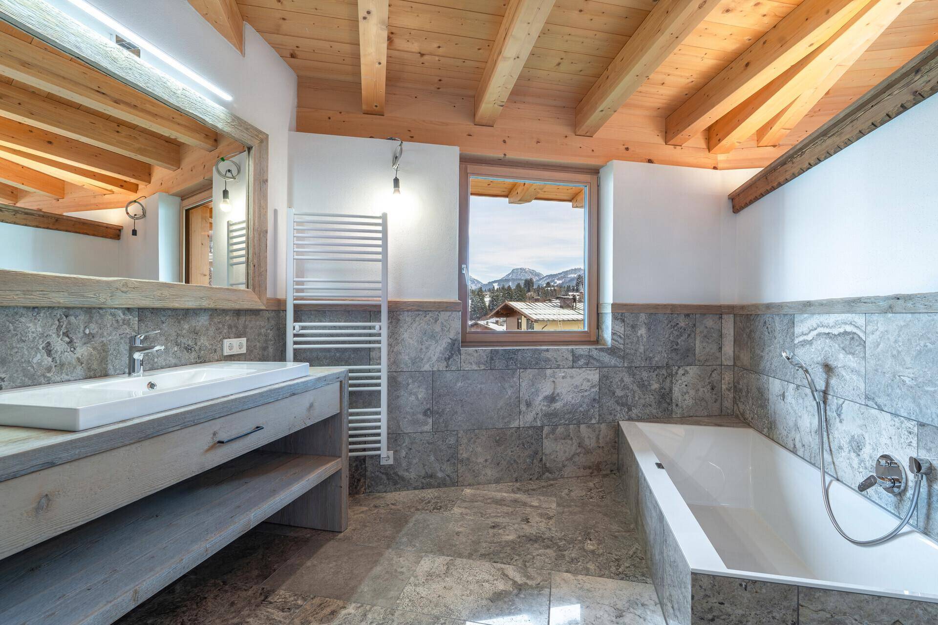 Modernes Badezimmer mit Badewanne, Einzelwaschbecken, grauen Fliesen und Bergblick aus dem Fenster.