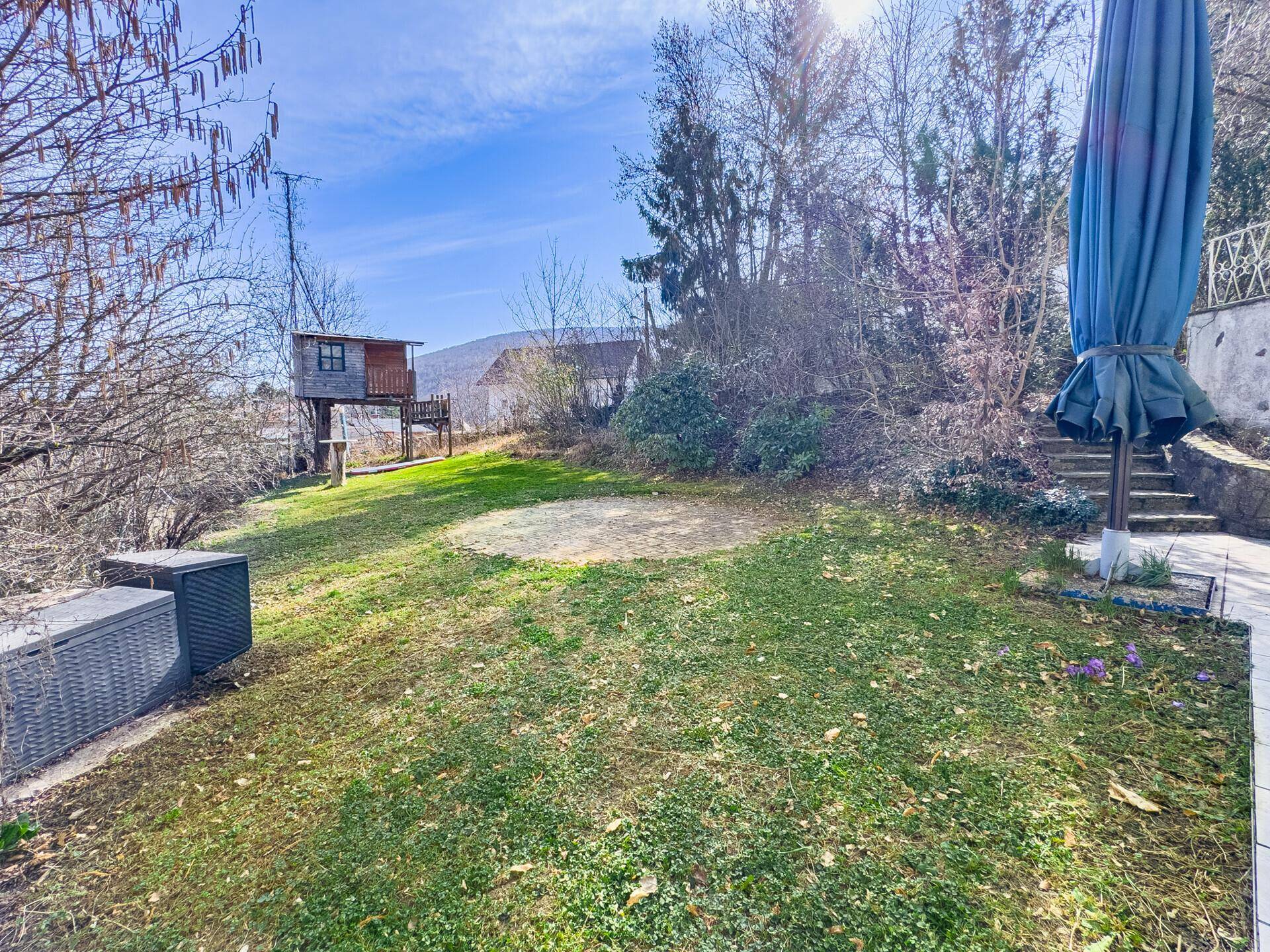 Gepflegter Garten mit Rasenfläche, Sitzgelegenheit und einem Baumhaus im Hintergrund.