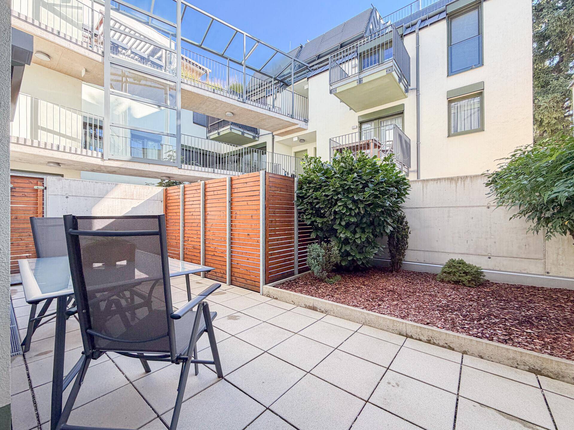 Gemütliche Terrasse mit Sitzgelegenheiten und kleinem Gartenbereich vor einem modernen Gebäude.