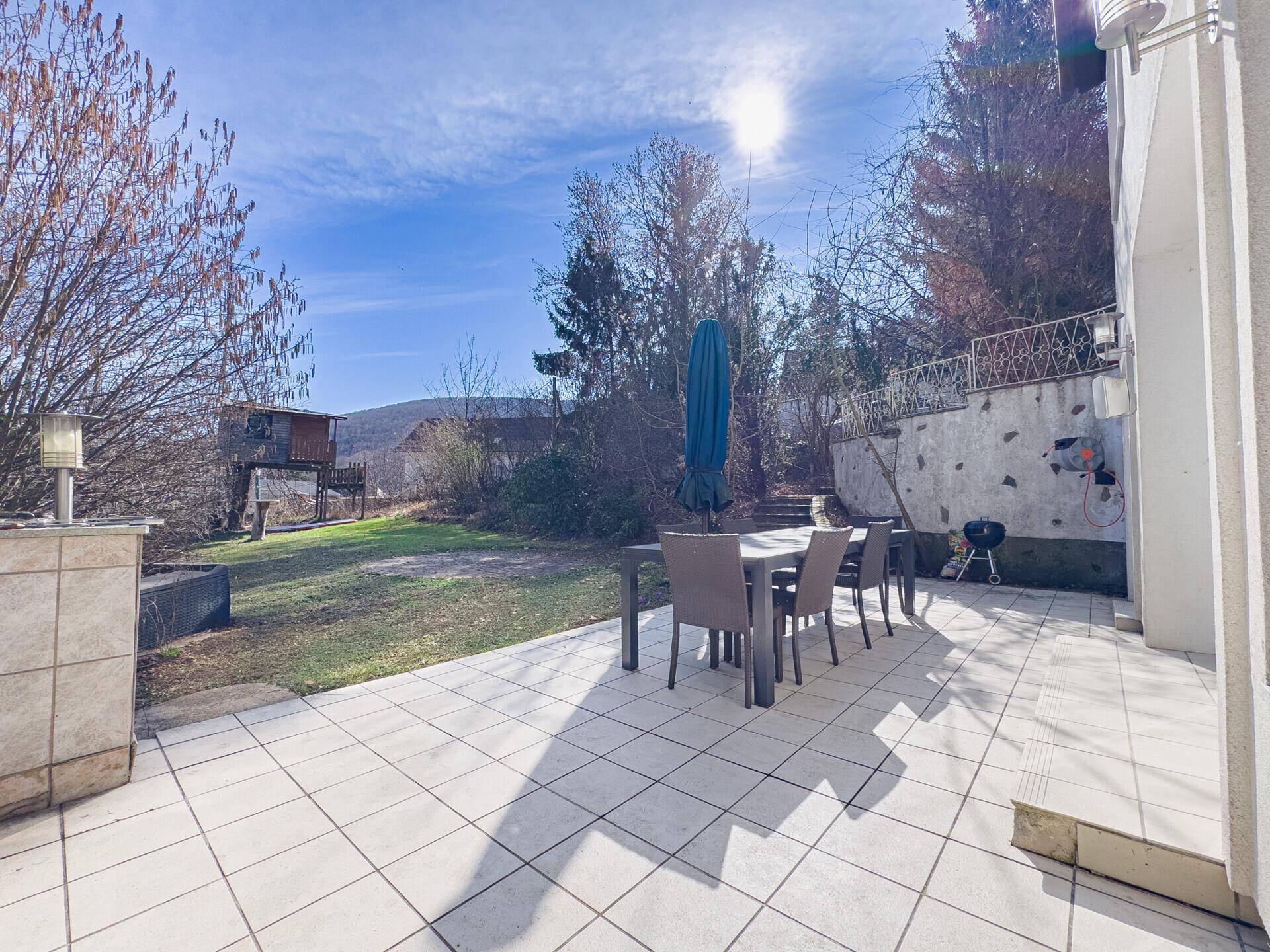 Sonniger Terrassenbereich mit Esstisch, Grill und Blick auf den angrenzenden Garten.