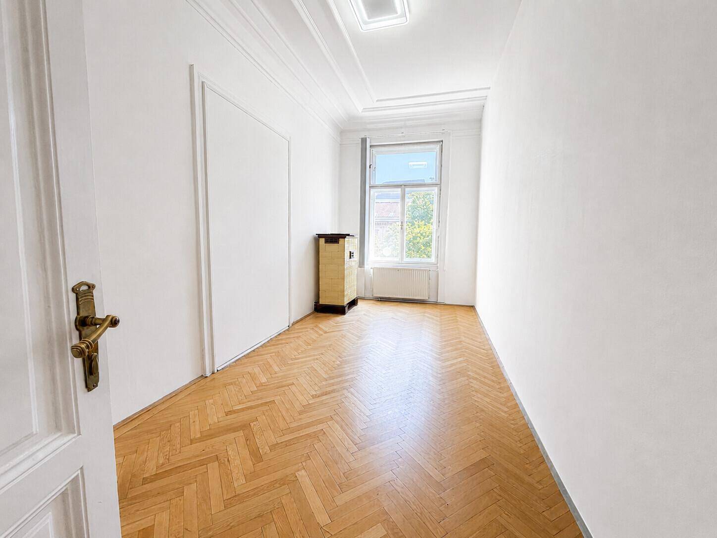 Langes, helles Zimmer mit Fischgrätparkett und einem Fenster, das viel Tageslicht hereinlässt.