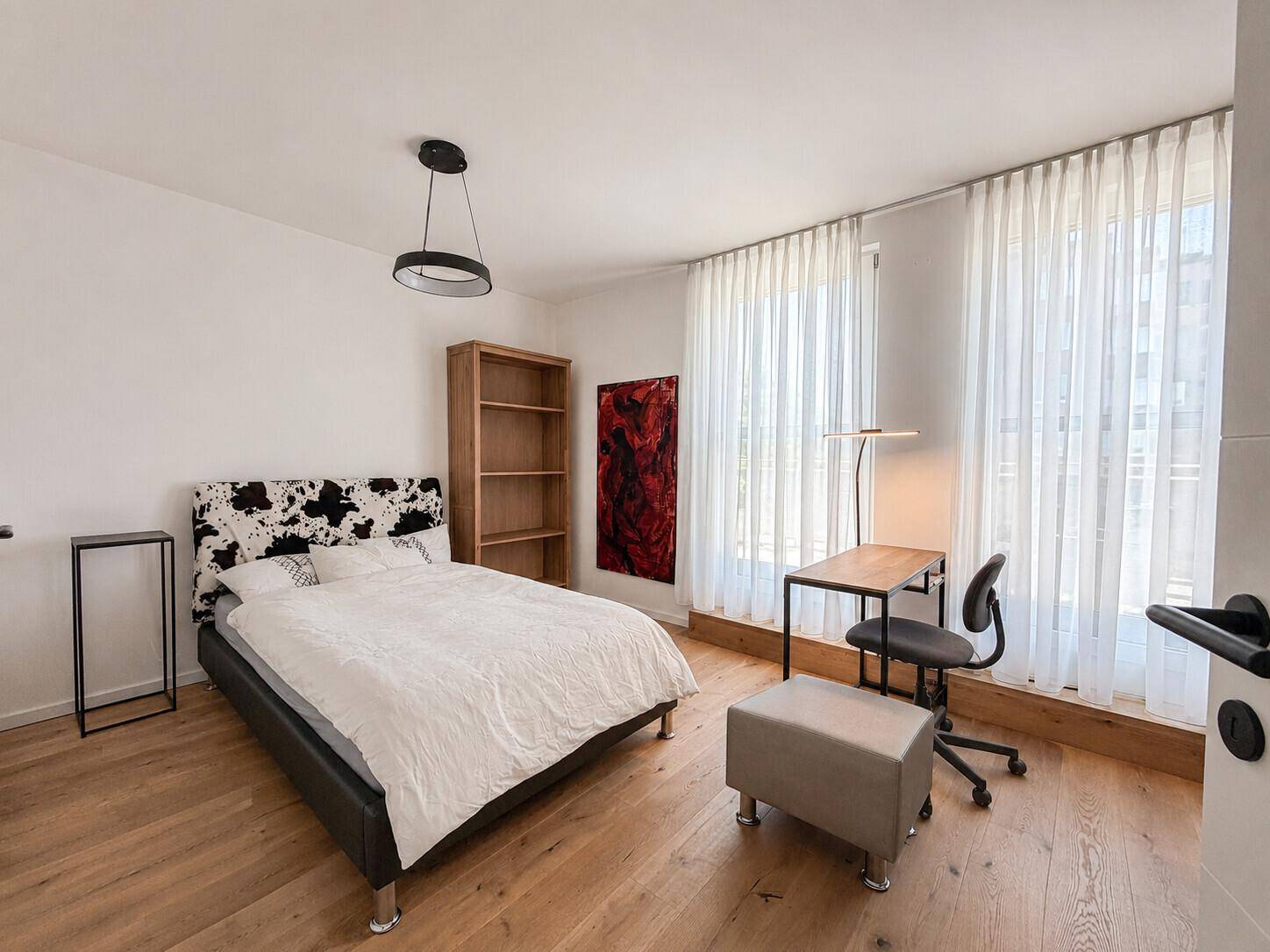 Geräumiges Zimmer mit Holzboden, Schreibtisch und großen Fenstern.